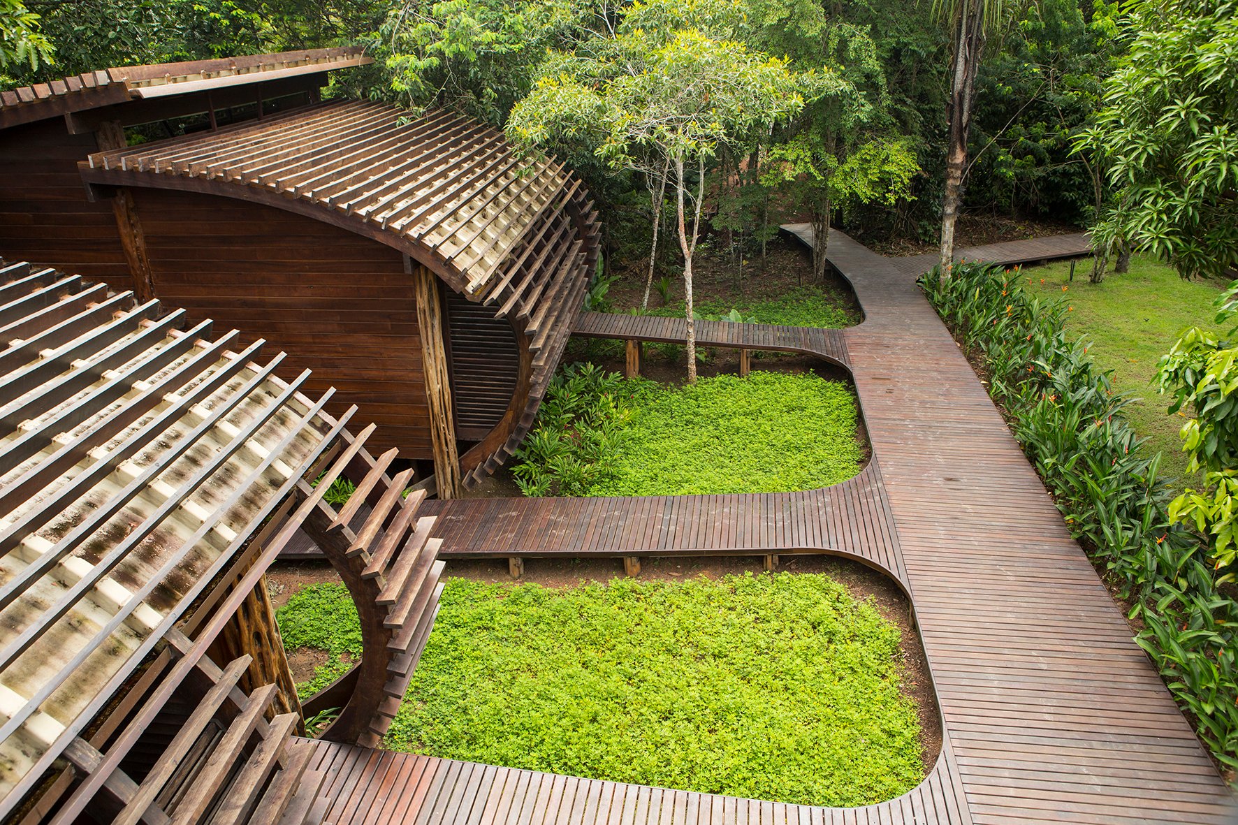 Hier buigt de architectuur mee met het woud. Bovenaanzicht van de slingerende hardhouten vlonders van Mirante do Gavião die kronkelen tussen weelderige groene bodembeplanting en een houten lamellenpaviljoen, met het dichte regenwoudkronendak op de achtergrond