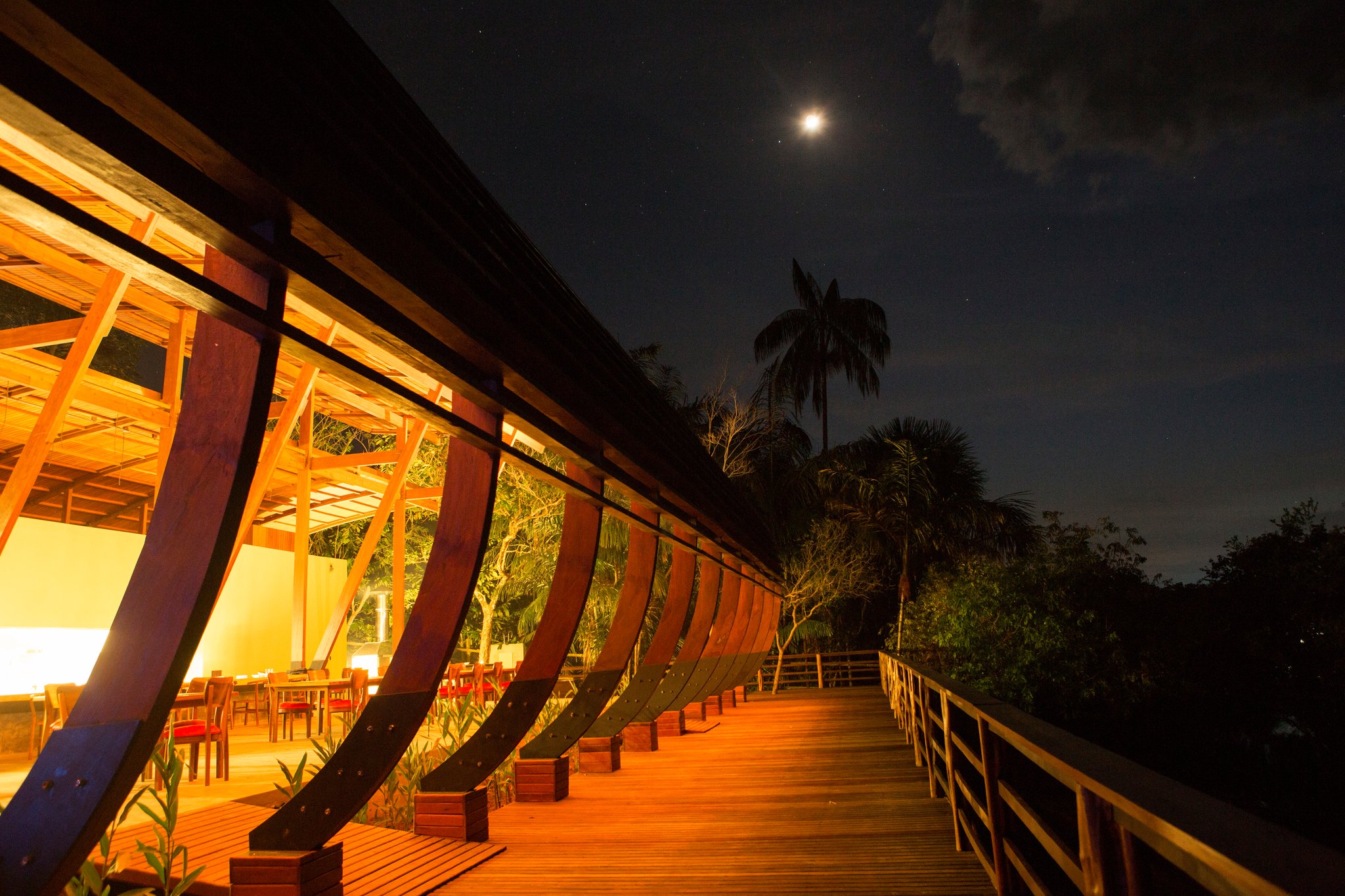 Het gebogen houten terras en de verlichte open eetpaviljoen van Mirante do Gavião onder een maanverlichte Amazone-nachtlucht, met palmboomsilhouetten tegen de sterrenhemel