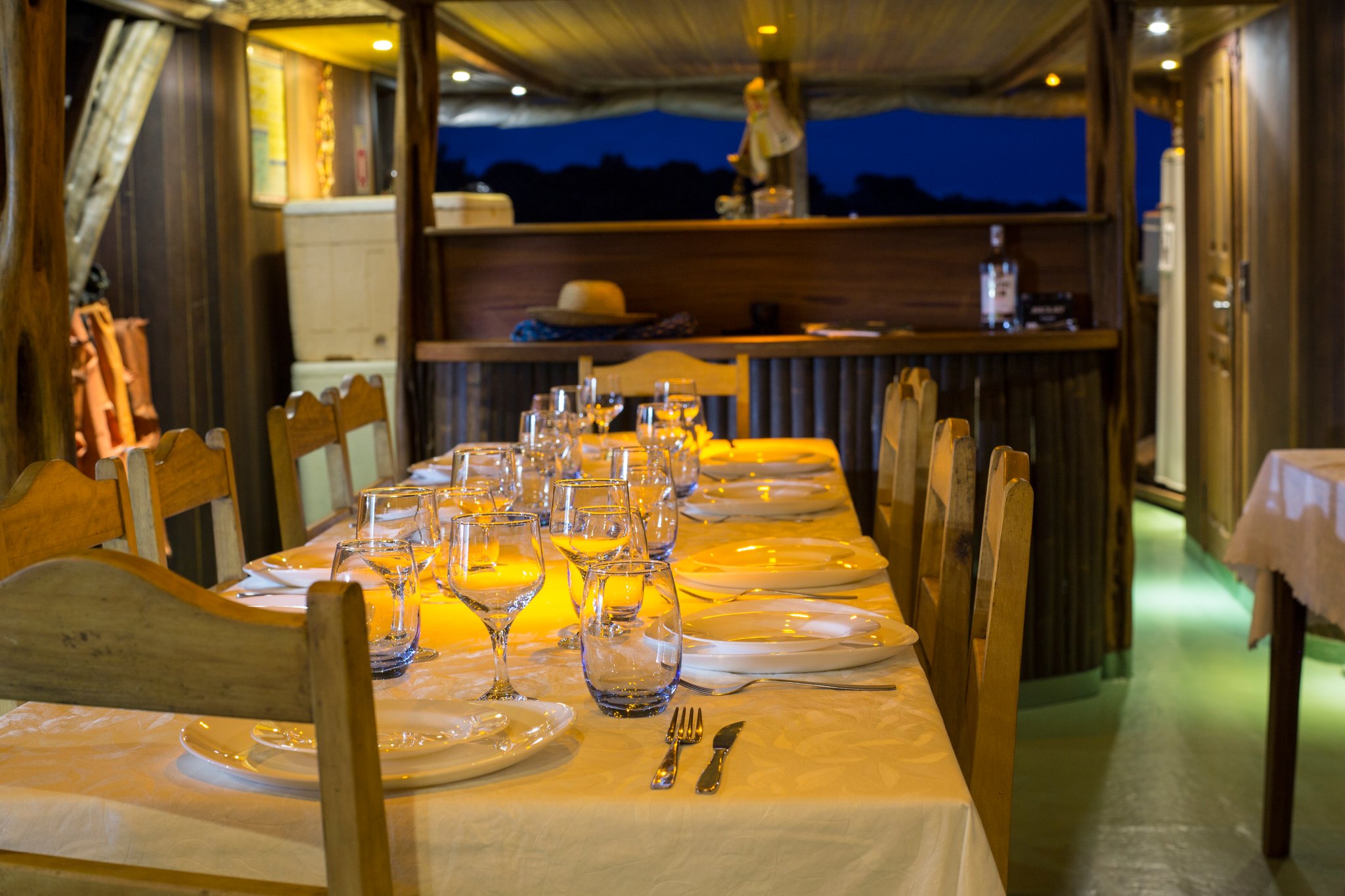 Dinner is ready, and the river is still out there waiting. A long dining table set with white linens, wine glasses and porcelain plates glowing under warm amber light aboard an Amazon jungle cruise Brazil riverboat at dusk