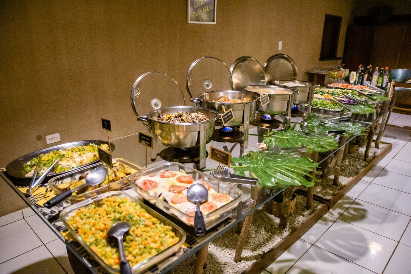 Een lang buffet in een budgetvriendelijke Pantanal-lodge met chafing dishes vol warm eten, een ovenschotel met tomaten, gehakte groentesalade, groene salades en tropische bladversieringen
