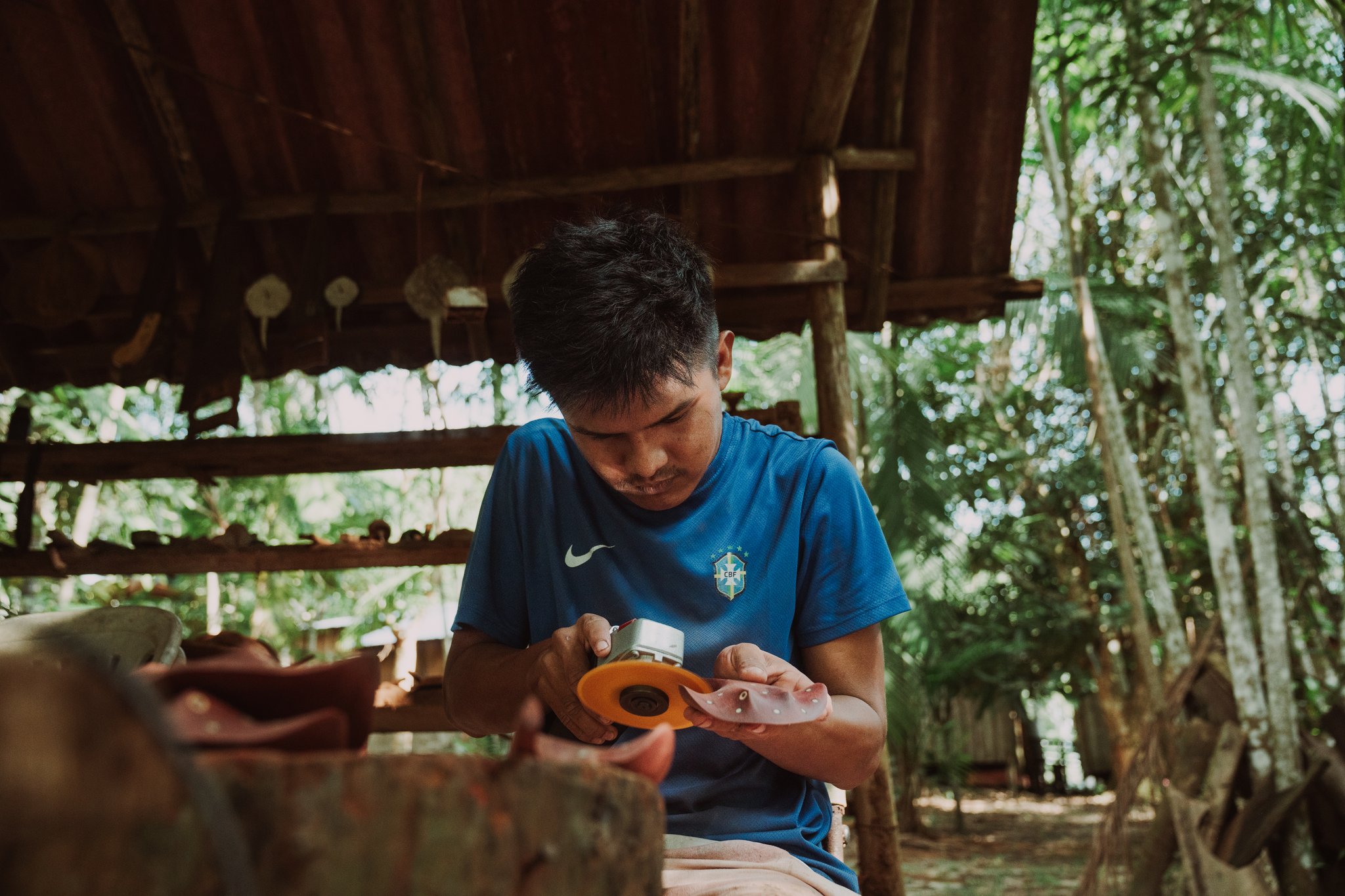 A man seated under a rustic palm-thatched open shelter carefully applies tape to a small handcrafted object, part of a community based tourism artisan workshop surrounded by lush tropical greenery