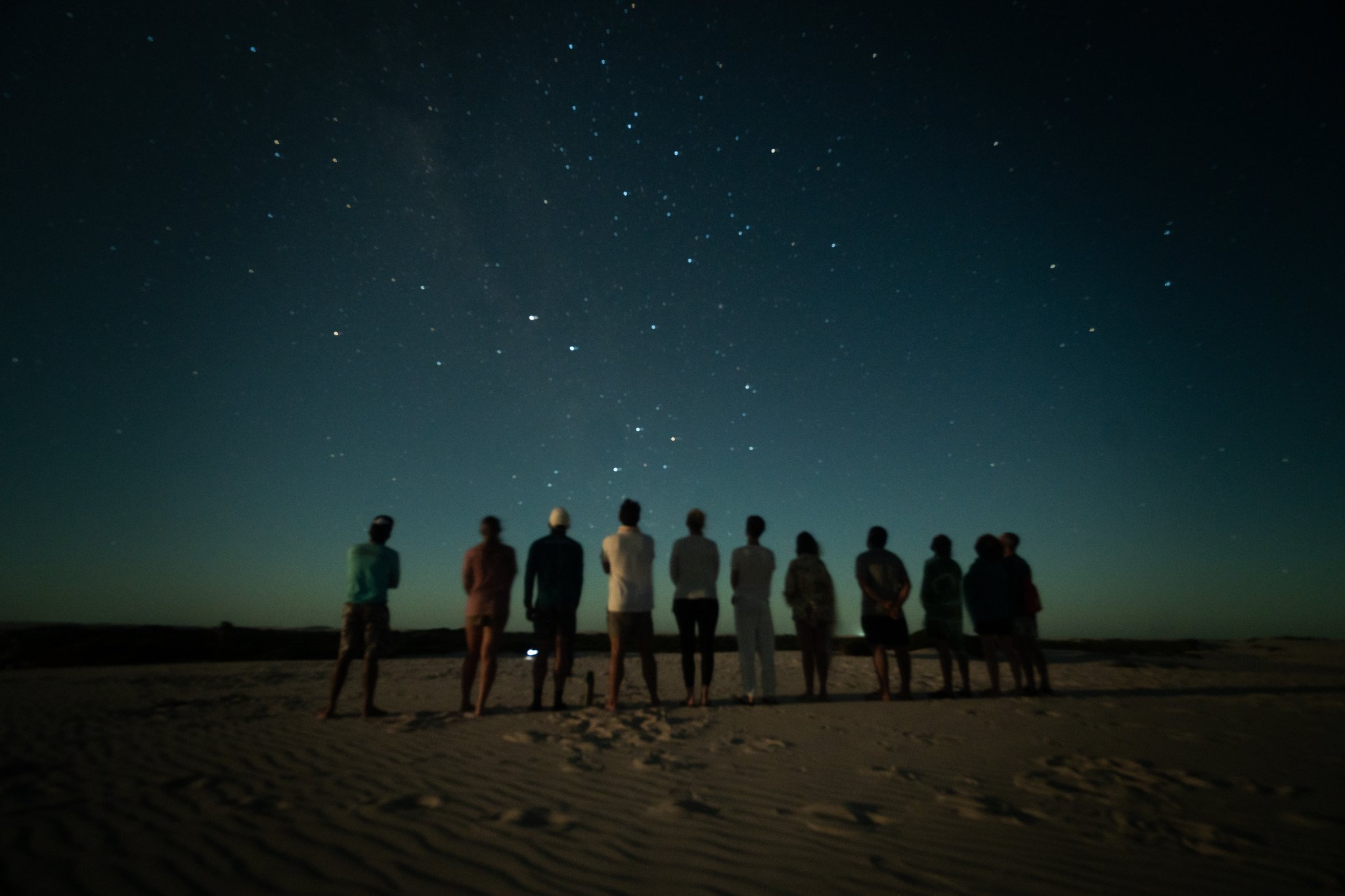 Zehn Menschen, kein Kunstlicht, und ein Himmel, der alles sagt. Eine Gruppe von Reisenden steht in einer Reihe auf weißen Sanddünen beim Lençóis-Maranhenses-Trek ab Atins, als Silhouetten vor einem tiefen blauen Nachthimmel voller Sterne und einem schwachen Milchstraßenband