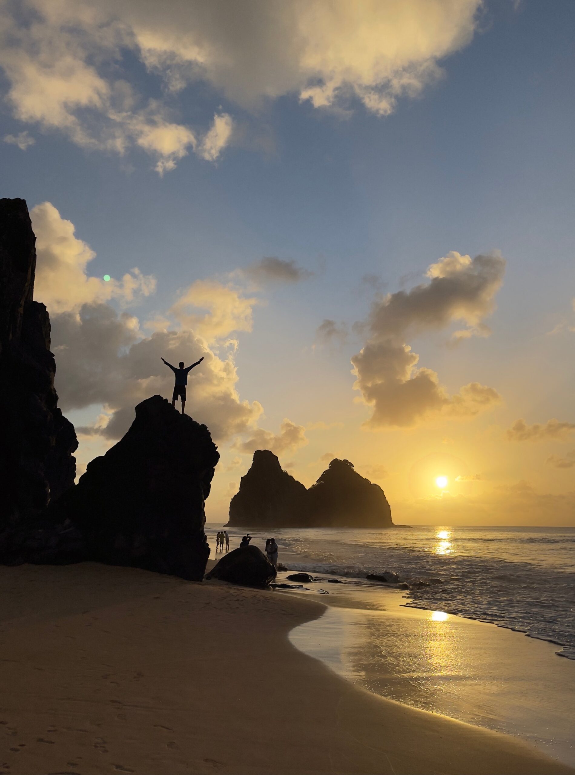 Uma pessoa de braços abertos em cima de uma rocha litorânea ao pôr do sol em Fernando de Noronha, com os picos do Morro Dois Irmãos em silhueta contra o céu dourado — o tipo de entardecer que justifica fazer um curso Open Water aqui.