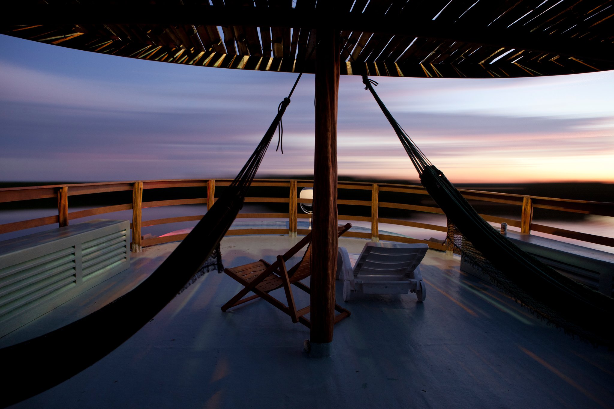 The river decides the schedule, and tonight it says: hammock. Hammocks and a wooden deck chair on the open upper deck of an Amazon jungle cruise Brazil vessel, framed by a thatched canopy, with a sweeping twilight sky and flat riverine horizon beyond the wooden railings