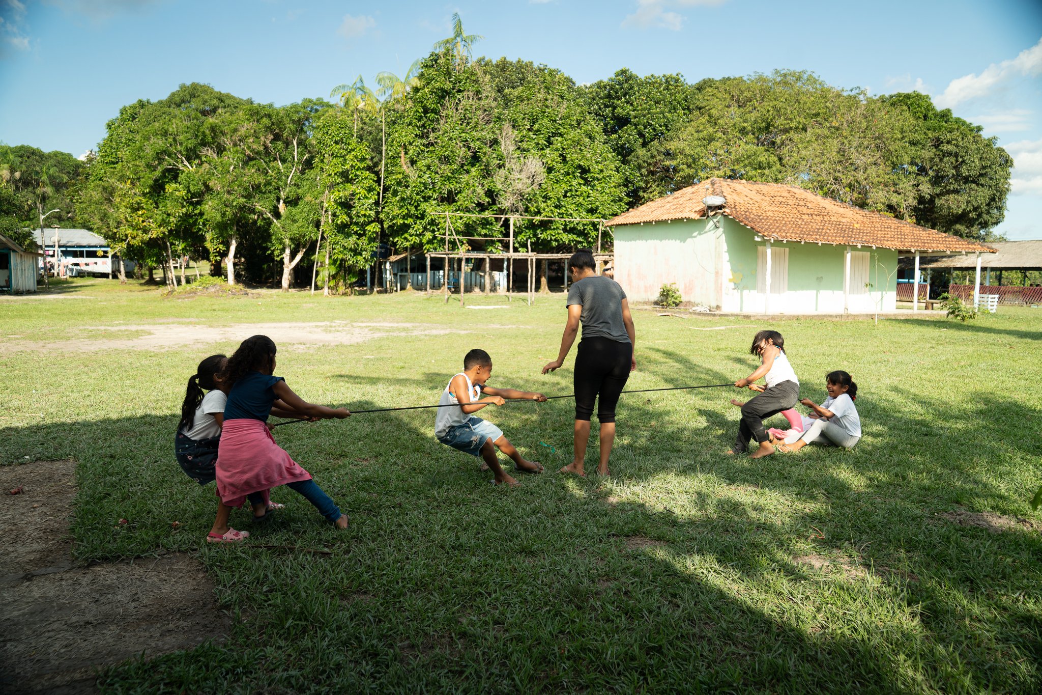 Children and adults playing tug-of-war on a grassy village green, with a terracotta-roofed house and tall tropical trees behind them — a candid moment of community based tourism where visitors are guests, not spectators