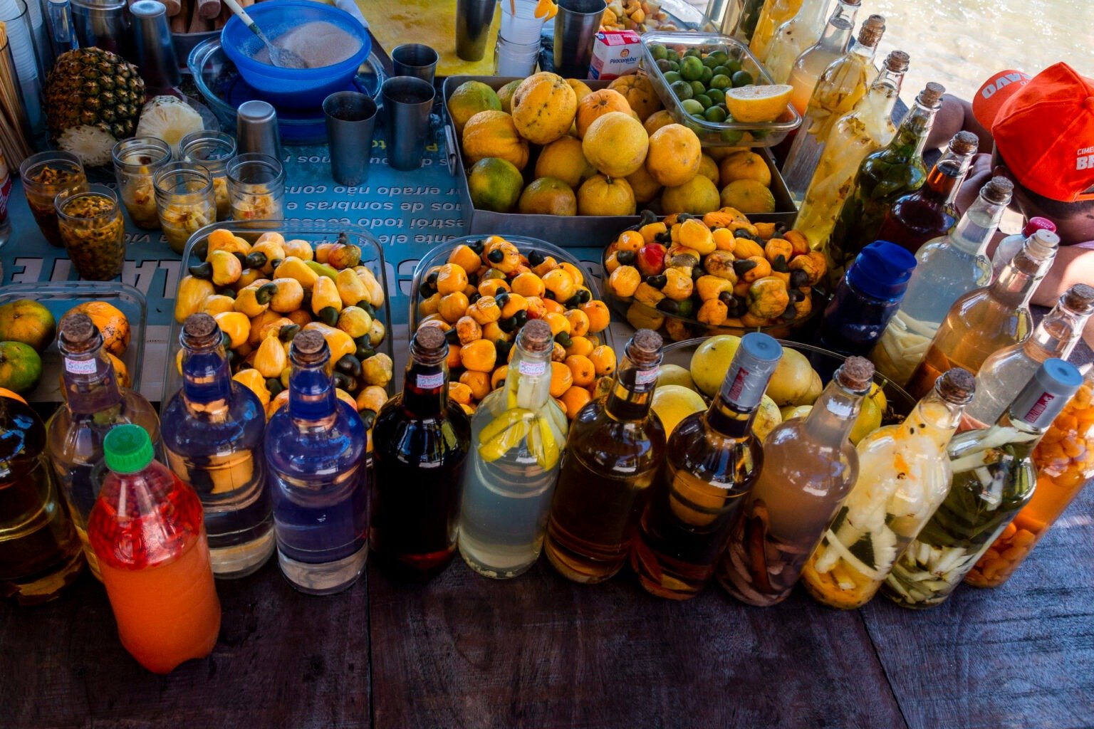 A market stall loaded with trays of fresh cashew fruit and tropical citrus alongside rows of colourful homemade infused liqueur bottles, part of the local food culture along the Lençóis Maranhenses full itinerary