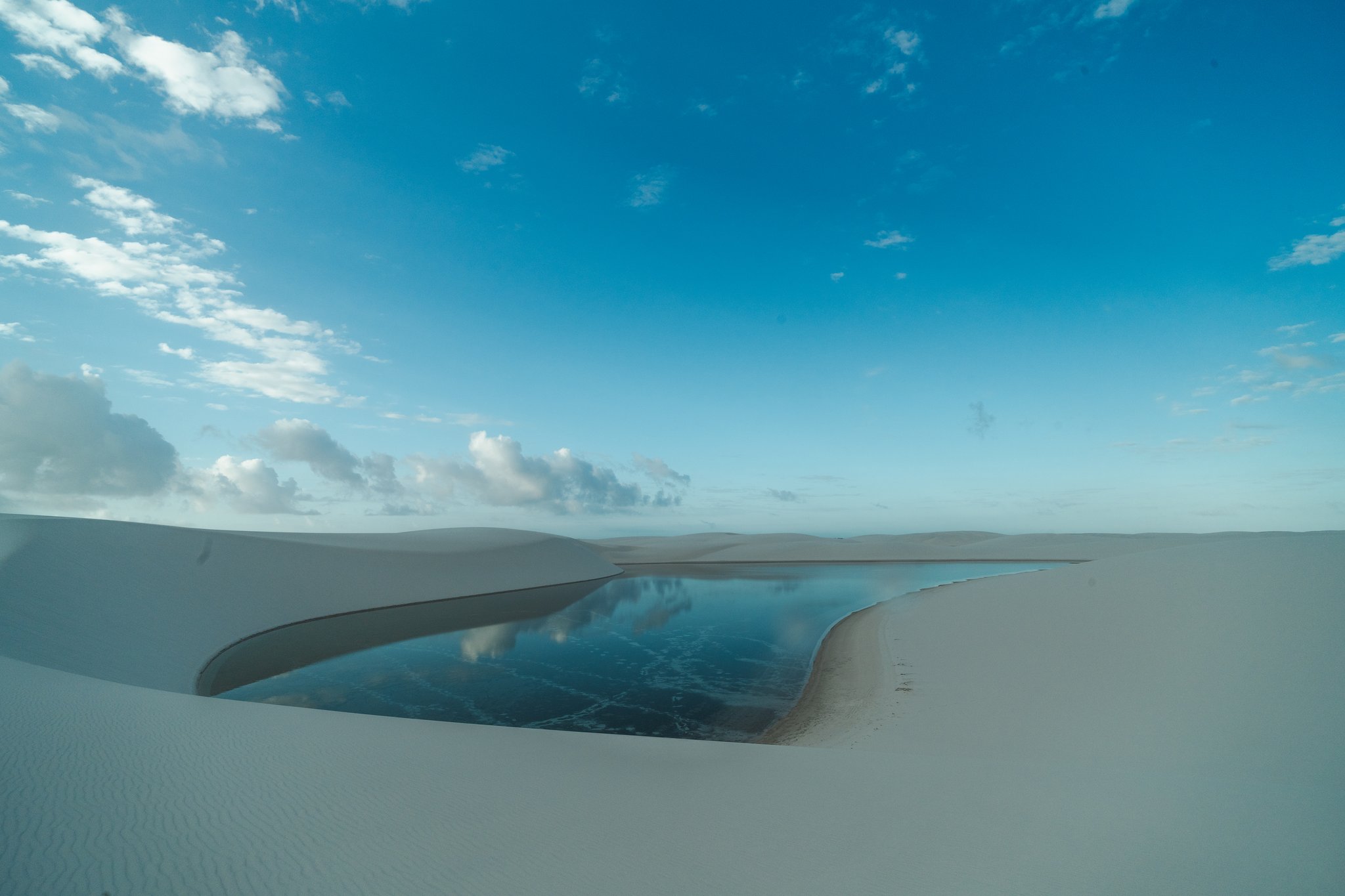 A still rainwater lagoon nestled between smooth white sand dunes reflects clouds and blue sky in the Lençóis Maranhenses Full Itinerary landscape