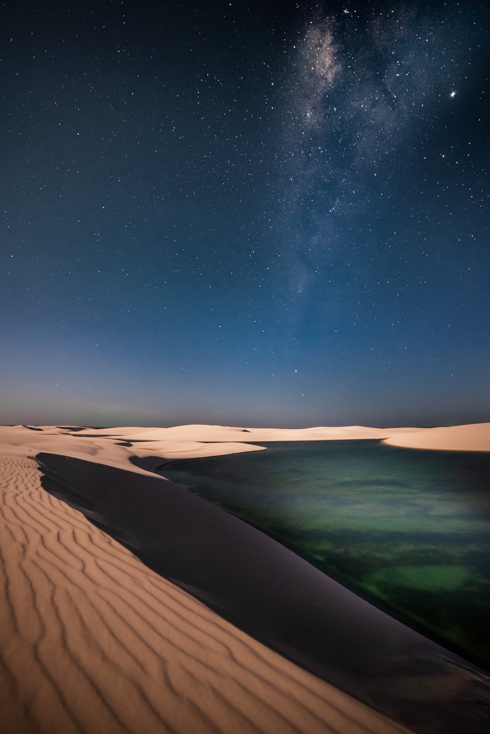 The Milky Way arching over white sand dunes and a still turquoise lagoon at night in Lençóis Maranhenses Full Itinerary stargazing experience