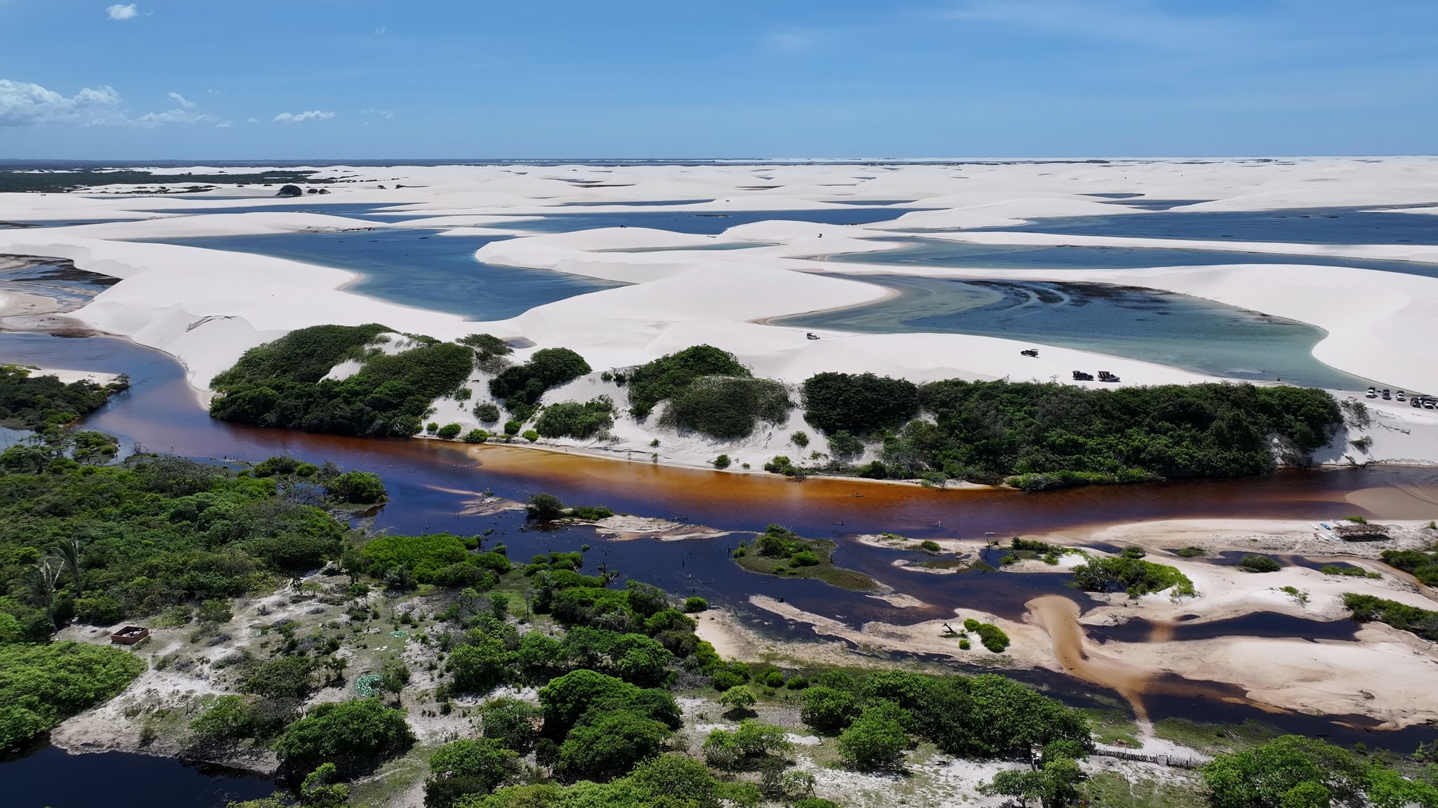 Aerial view of a tannin-dark river winding through green vegetation before meeting the white dunes and turquoise lagoons of the Lençóis Maranhenses full itinerary landscape