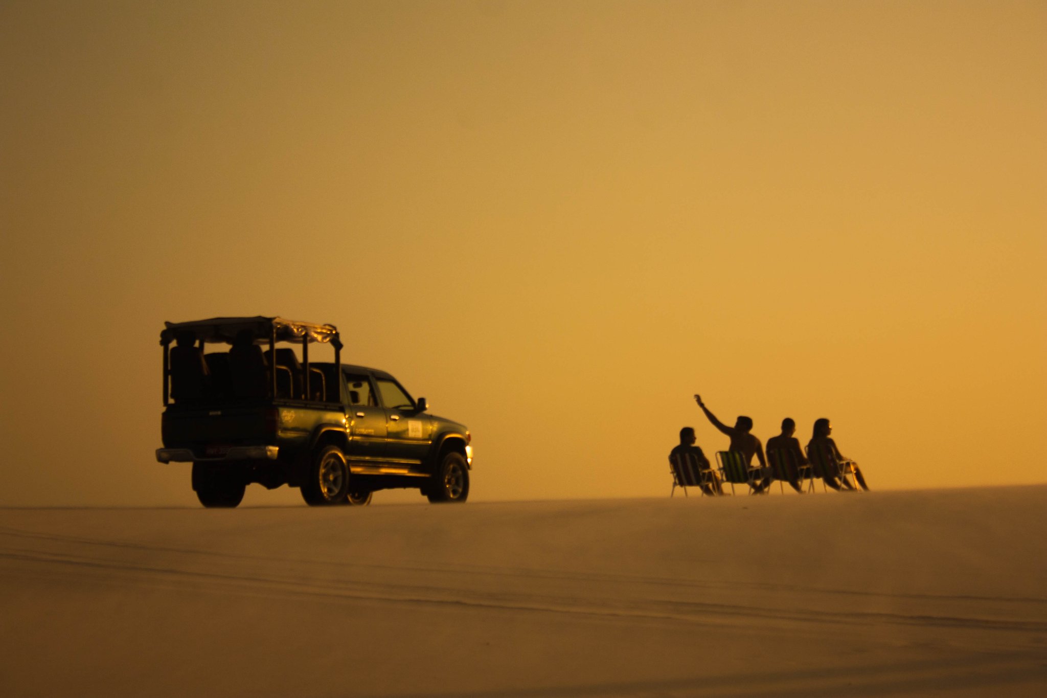 Four travelers seated in folding chairs on a vast open dune at golden-hour sunset, with a 4x4 safari vehicle silhouetted beside them during a Lençóis Maranhenses full itinerary excursion
