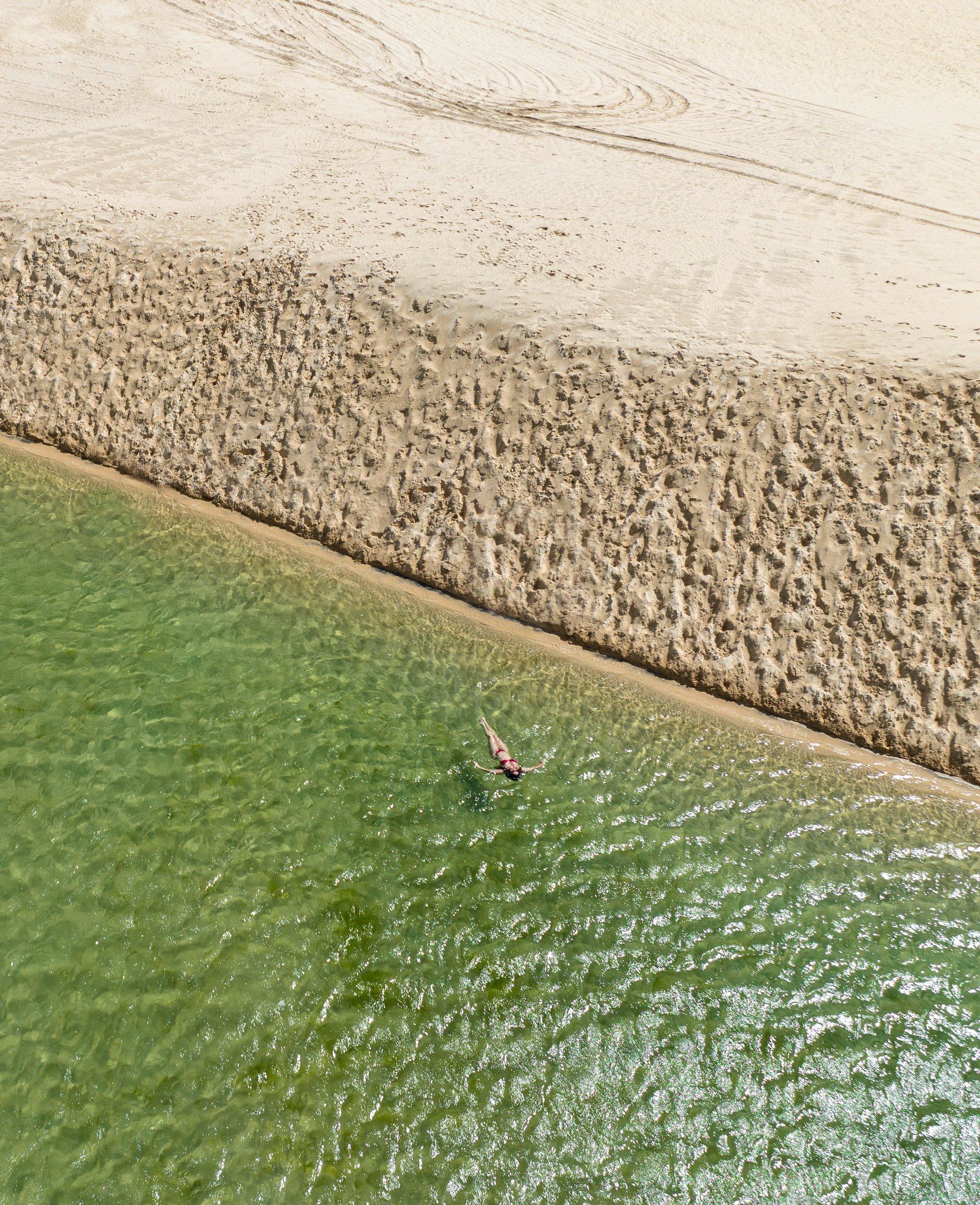 Ein Sandstreifen, grünes Wasser, und niemand, der einen stört. Luftaufnahme einer einzelnen Person, die im klaren grünen Wasser neben einer strukturierten Sandbank beim Lençóis-Maranhenses-Trek ab Atins schwimmt