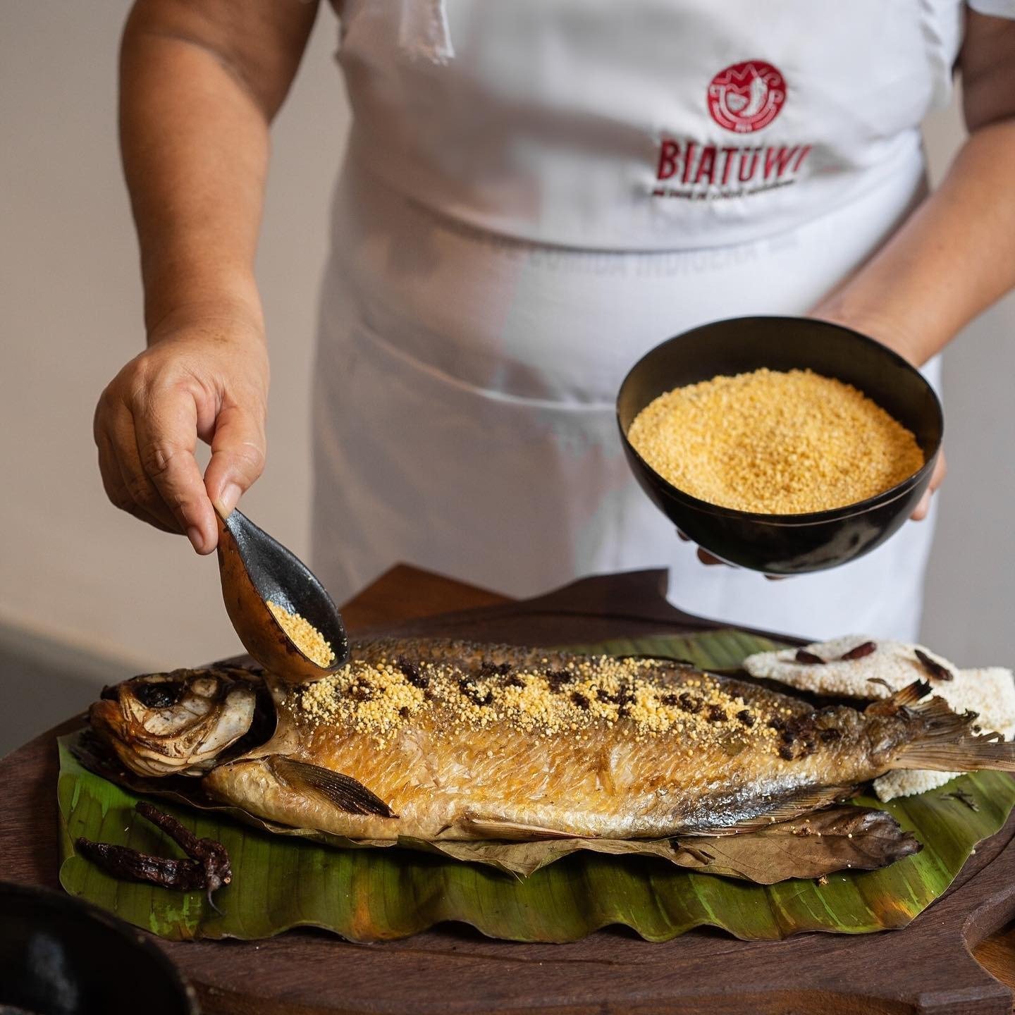 A cook wearing a Biatüwi apron spoons golden manioc farofa over a whole roasted fish laid on banana leaves, part of the regional cuisine served on an Amazon Cruise Trip in Brazil