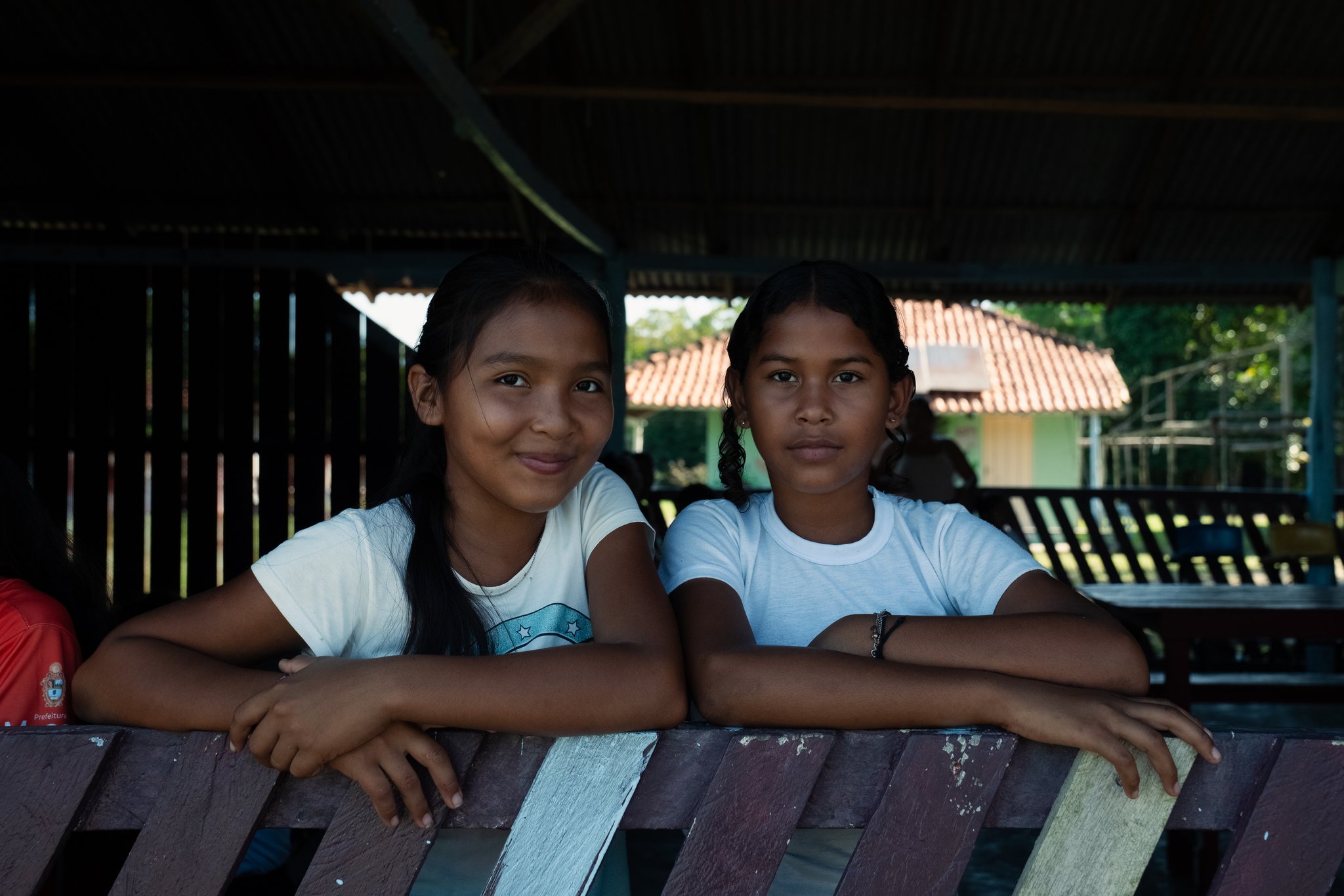 Two girls leaning over a wooden bench inside a covered community gathering space, smiling at the camera during a community based tourism visit in Brazil