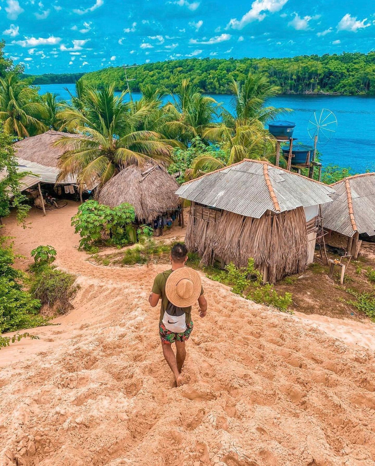 A traveller with a straw hat and backpack walks barefoot down a sandy slope toward thatched and tin-roofed huts nestled among palm trees beside a wide blue river on the Lençóis Maranhenses full itinerary