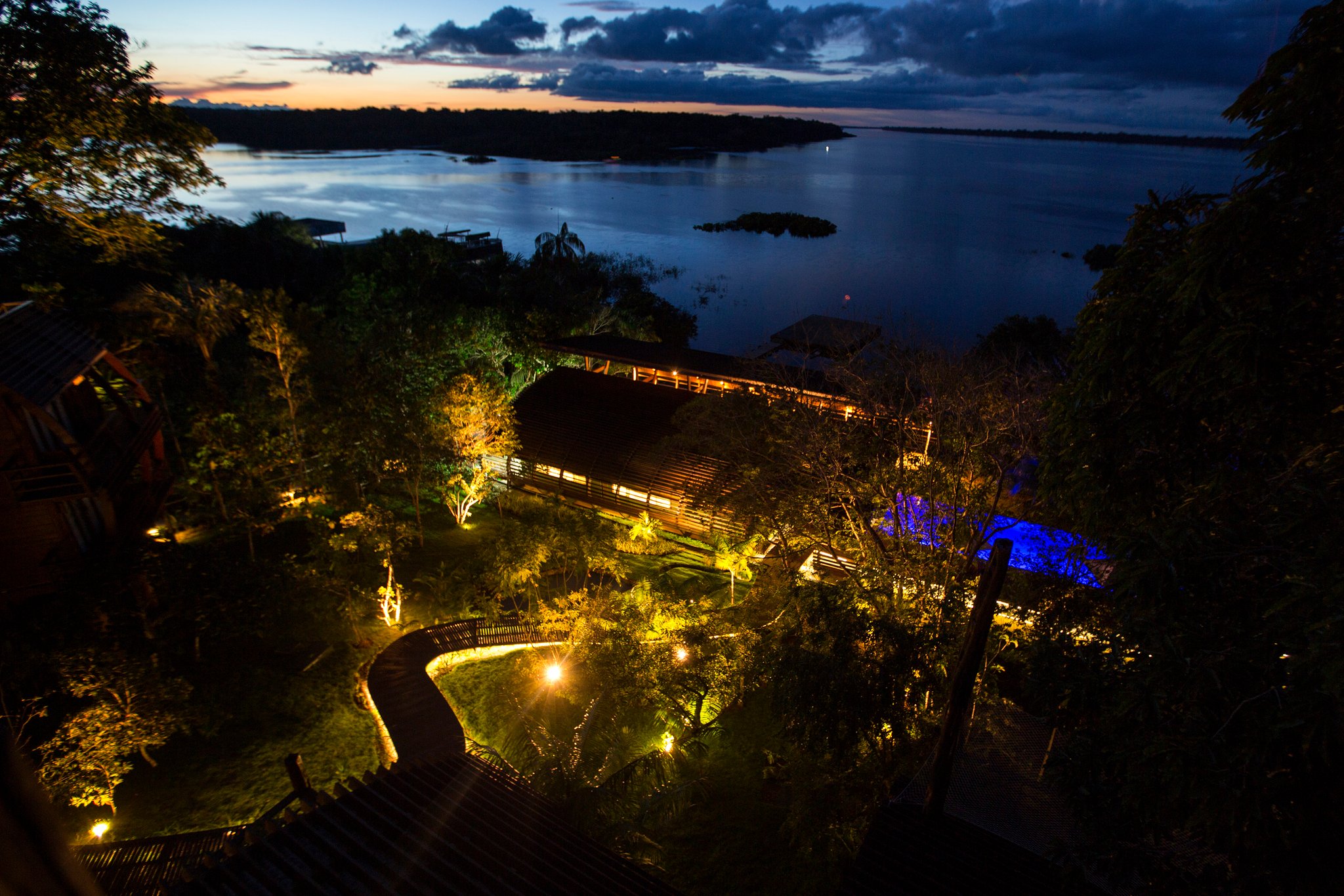 Op dit uur houdt het regenwoud even zijn adem in, en de lodge gloeit. Schemerfoto vanuit de lucht van Mirante do Gavião met warm tuinlicht, een kronkelend houten pad, een blauw verlicht zwembad en de brede Rio Negro onder een diepblauwe avondlucht
