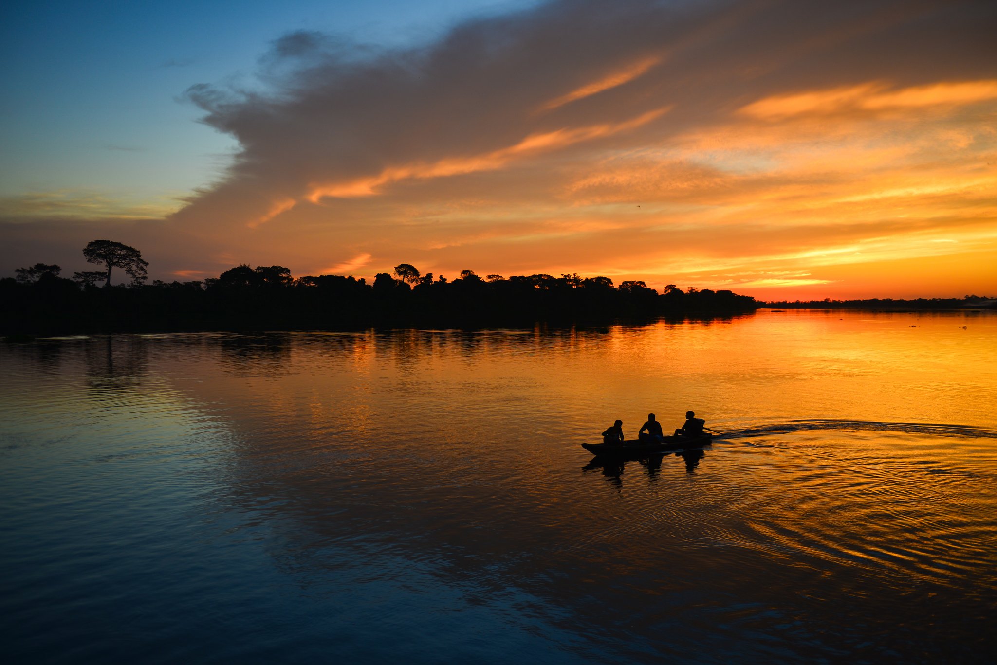 Three people silhouetted in a small wooden canoe crossing a wide river at sunset on a tucano amazon river cruise, orange and gold clouds reflected in the calm dark water, dense forest treeline on the horizon