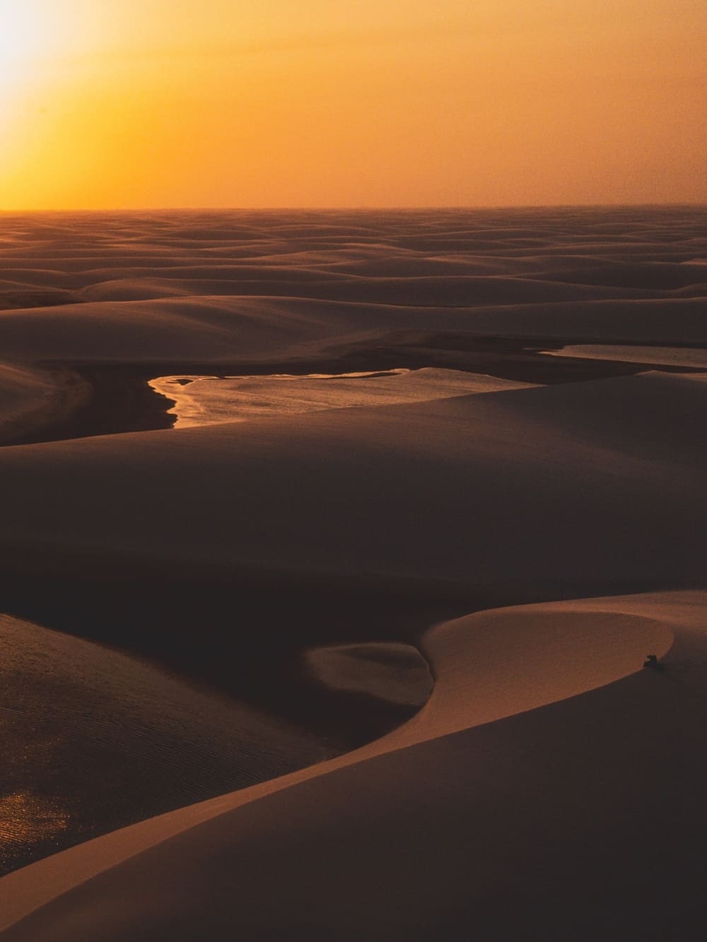 A vast expanse of smooth sand dunes illuminated by a vibrant golden sunset in Lençóis Maranhenses National Park, Brazil. A shallow pool of water rests in the valley between the dunes, reflecting the bright orange sky
