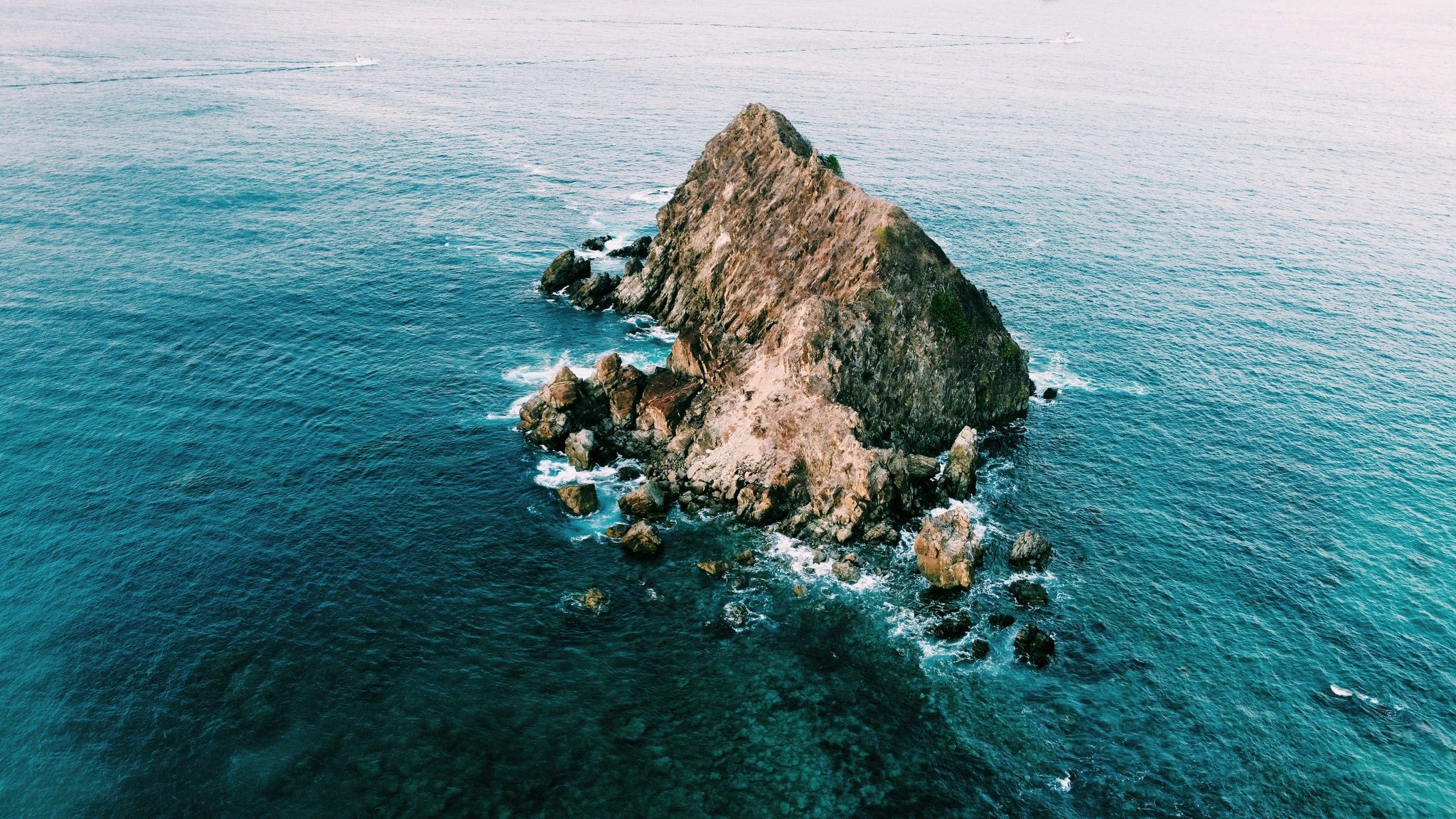 Aerial view of a jagged rocky islet rising from deep blue open ocean, surrounded by scattered boulders and white surf — a dramatic seascape near Fernando de Noronha on this 2-Day Diving Package in Fernando de Noronha