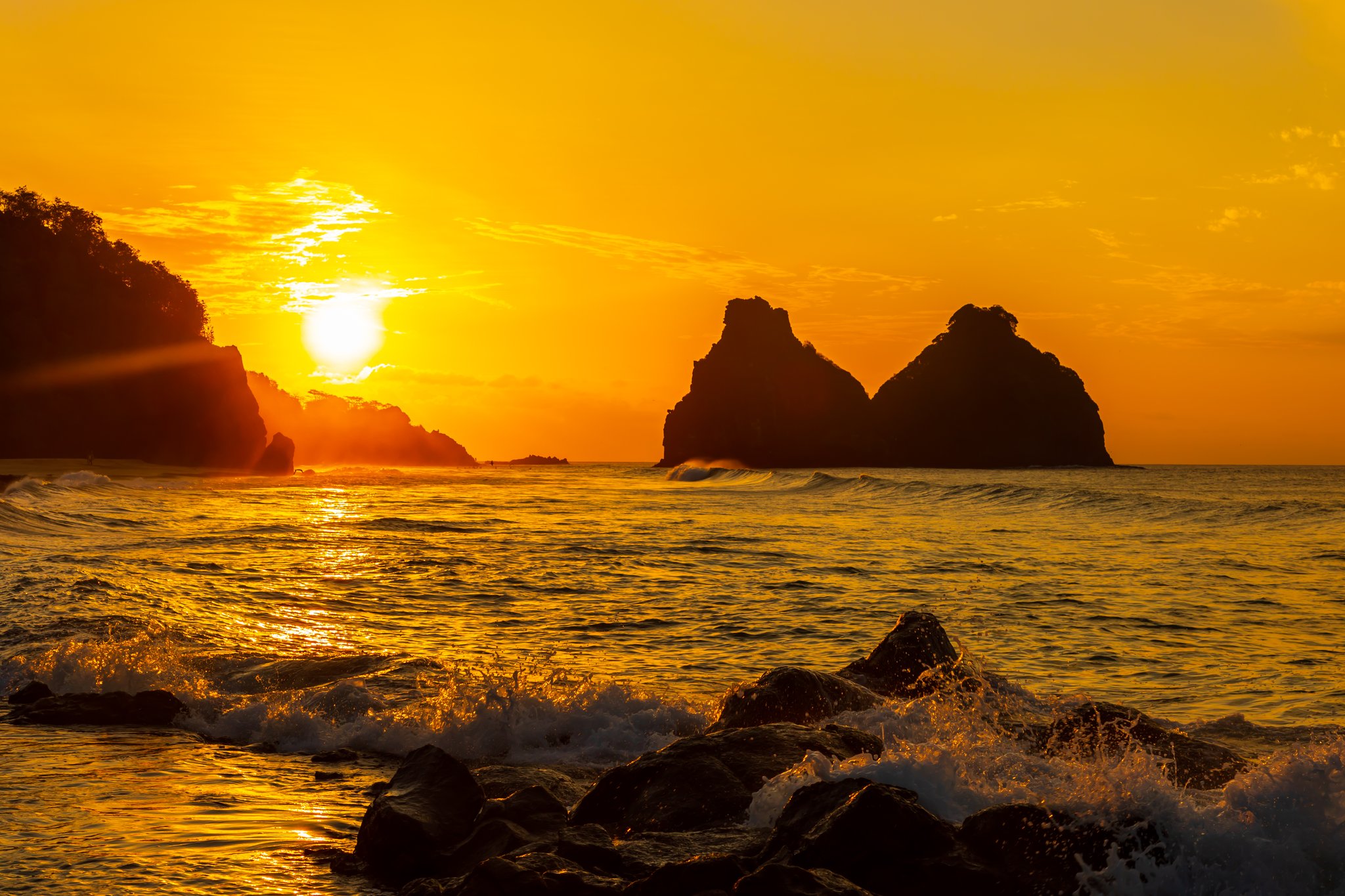 Fiery golden sunset over silhouetted rocky sea islets off Fernando de Noronha, waves crashing over foreground boulders, part of a 2-Day Diving Package in Fernando de Noronha