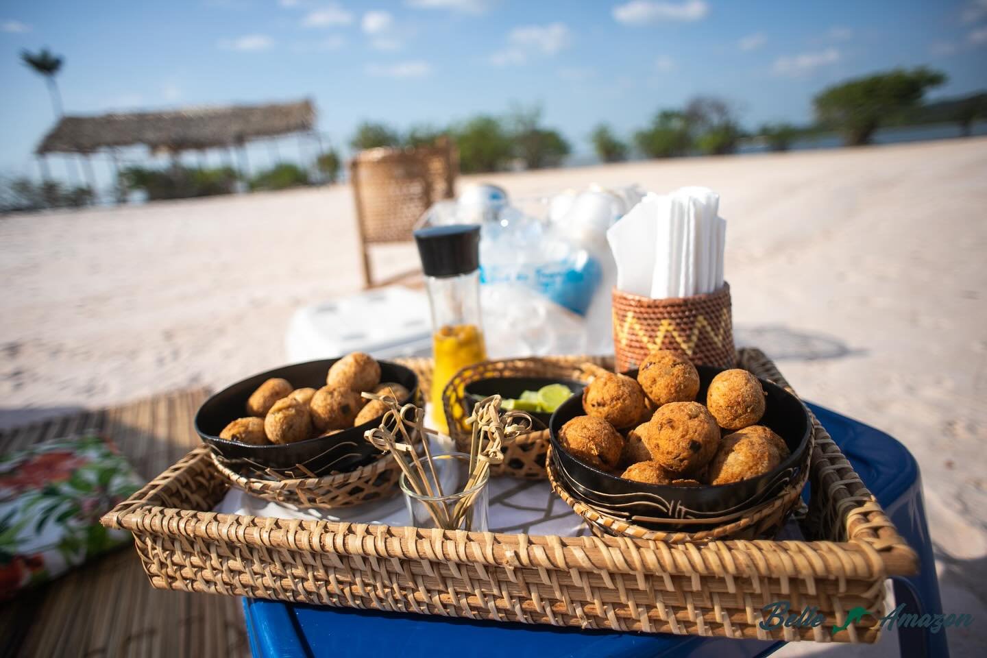 Golden bites, river sand, open sky — snack time on the Tapajós. A woven rattan tray holding two black bowls of golden fried snack balls served on a sandy riverbank during an Amazon cruise from Alter do Chão, with bamboo skewers, lime wedges and condiments alongside