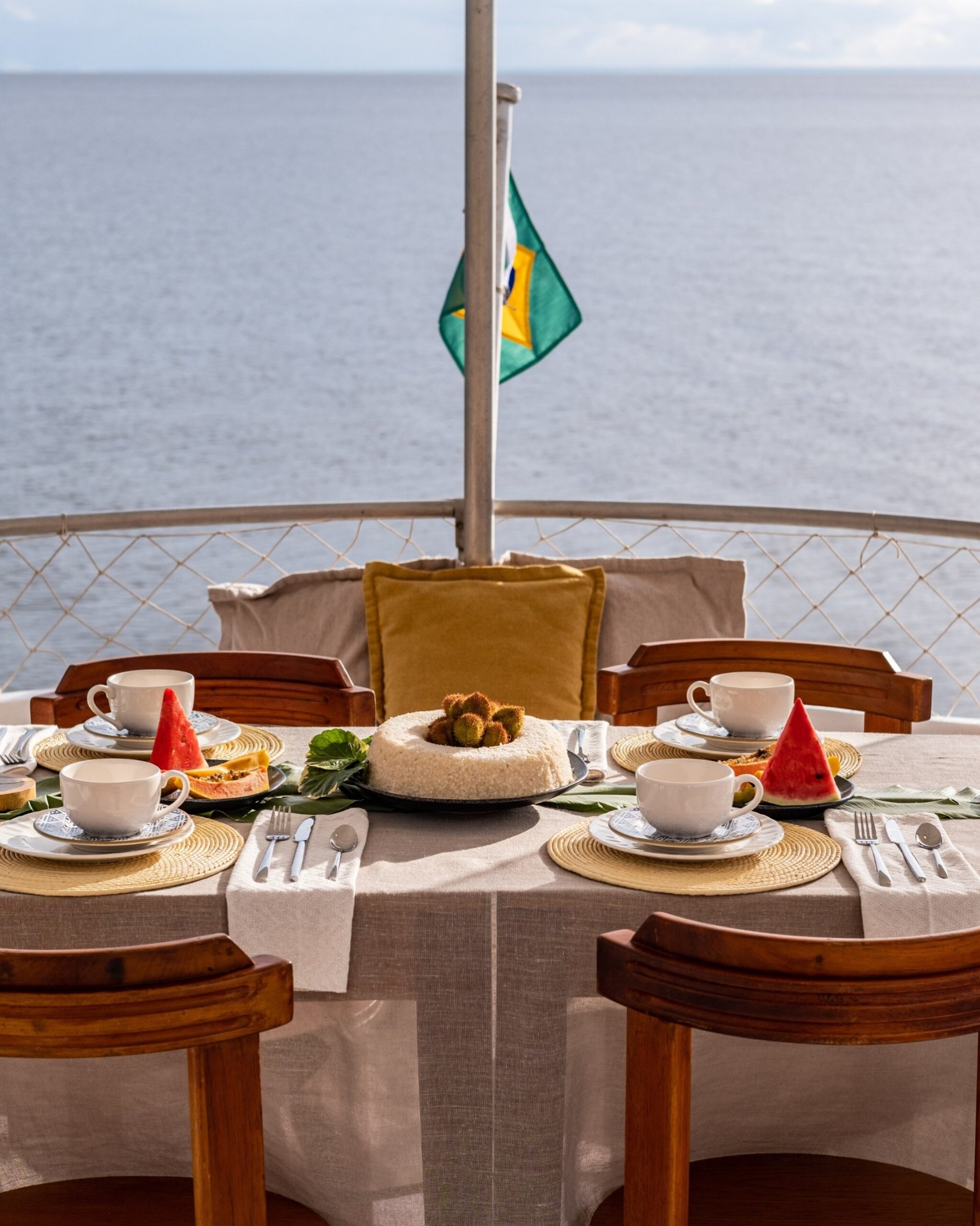 Breakfast laid, river stretching to the horizon — mornings on the water taste different. A set breakfast table on the open deck of an Amazon cruise from Alter do Chão, with fresh watermelon, papaya, white coffee cups, and a centrepiece cake, a Brazilian flag flying over calm open water in the background