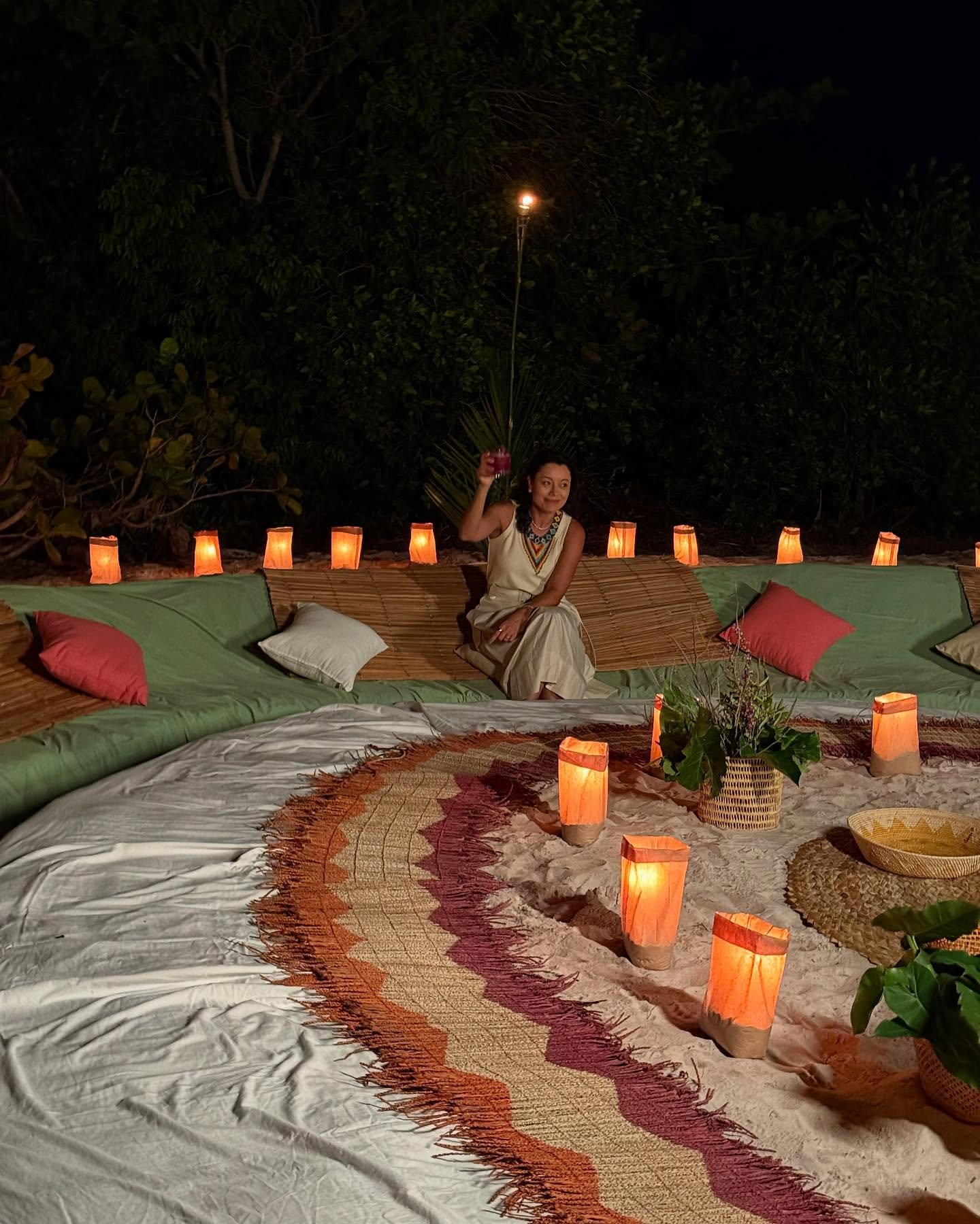 Manchmal braucht der Abend am Amazonas nur ein paar Laternen und gute Gesellschaft. Eine Frau in weißem Kleid kniet zwischen leuchtenden Papierlaternen und geflochtenen Matten auf einem Sandstrand bei Nacht, Teil eines Freiluftdinner-Setups auf einer Amazonas-Kreuzfahrt ab Alter do Chão