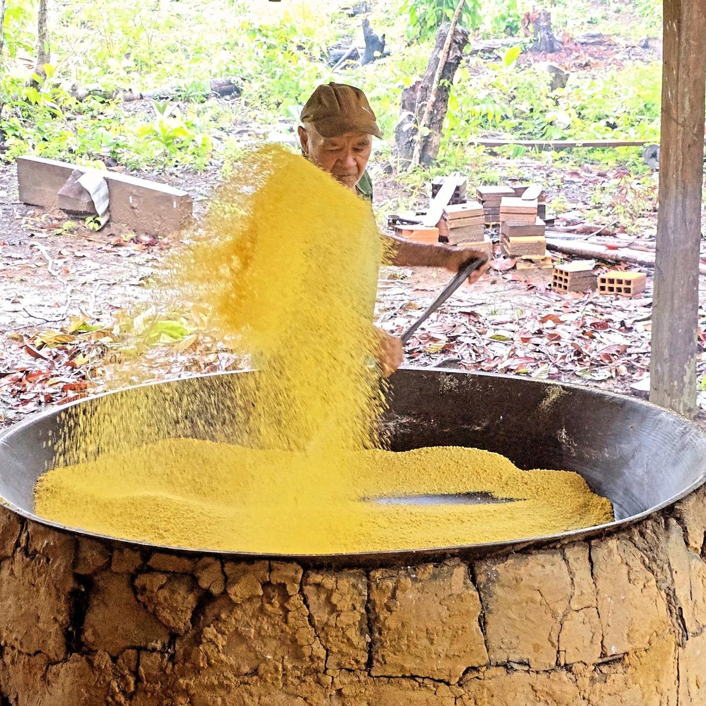 Flour flying, arms working — cassava transformed by hand the way it has been for generations. An elder man toasts golden cassava flour in a large iron pan over a clay wood-fired oven, part of a community visit on an Amazon cruise from Alter do Chão