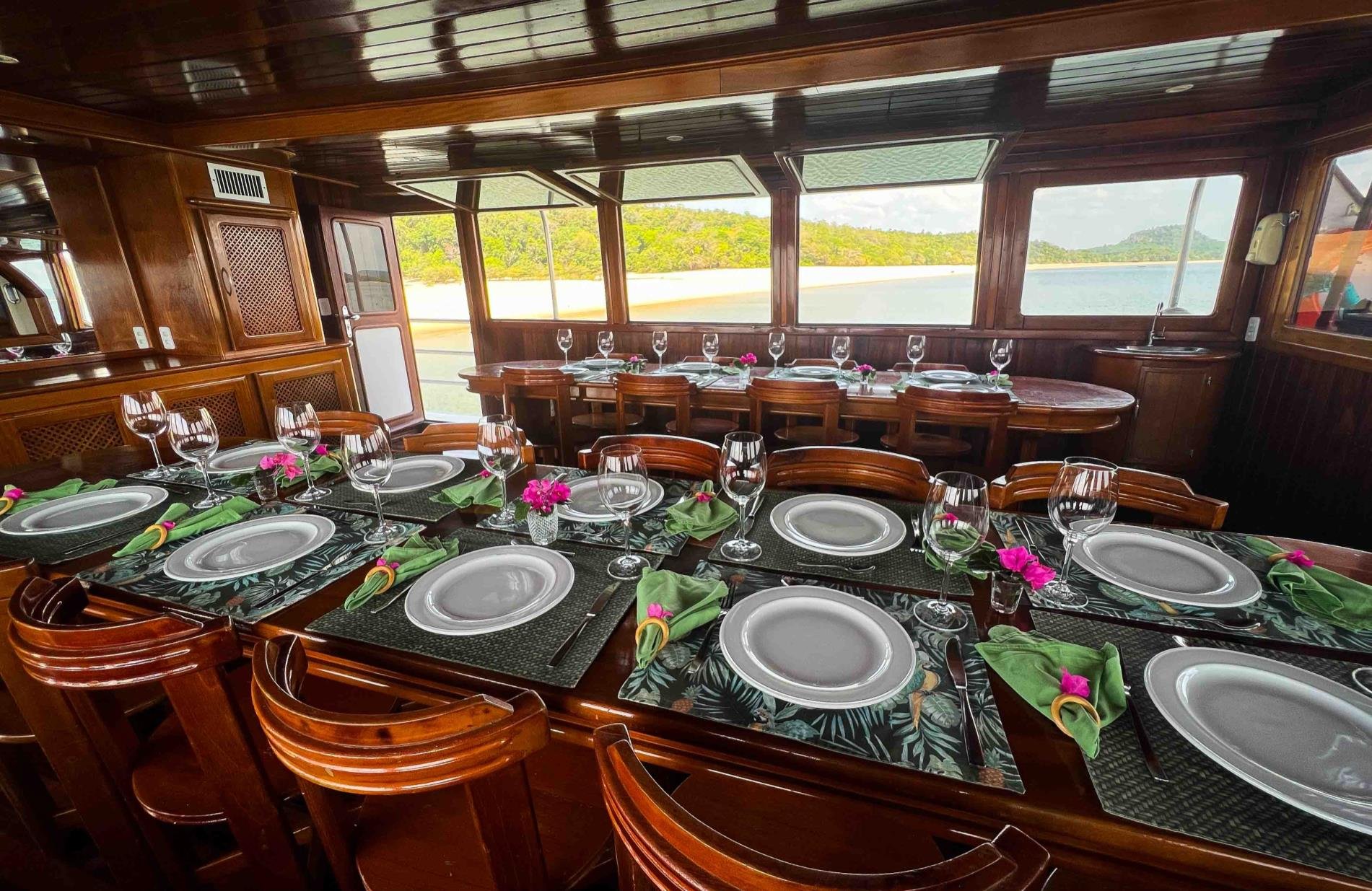 Abendessen mit Flussblick, so sieht ein guter Abend auf dem Amazonas aus. Elegant gedeckte Esstische mit Weingläsern, grünen Servietten und rosa Blütendekor im polierten Holzsalon einer Amazon-Kreuzfahrt von Alter do Chão, mit Blick auf einen Flussstrand und bewaldete Hügel durch Panoramafenster