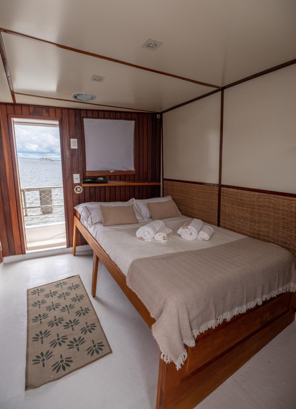 Schlafen, während der Amazonas vorbeizieht, das klingt besser als jedes Hotel. Ein ordentlich gemachtes Doppelbett mit gerollten weißen Handtüchern in einer Kabine mit Rattankopfteil an Bord einer Amazonas-Kreuzfahrt, durch die offene Tür ist ein ruhiger Flussblick zu sehen