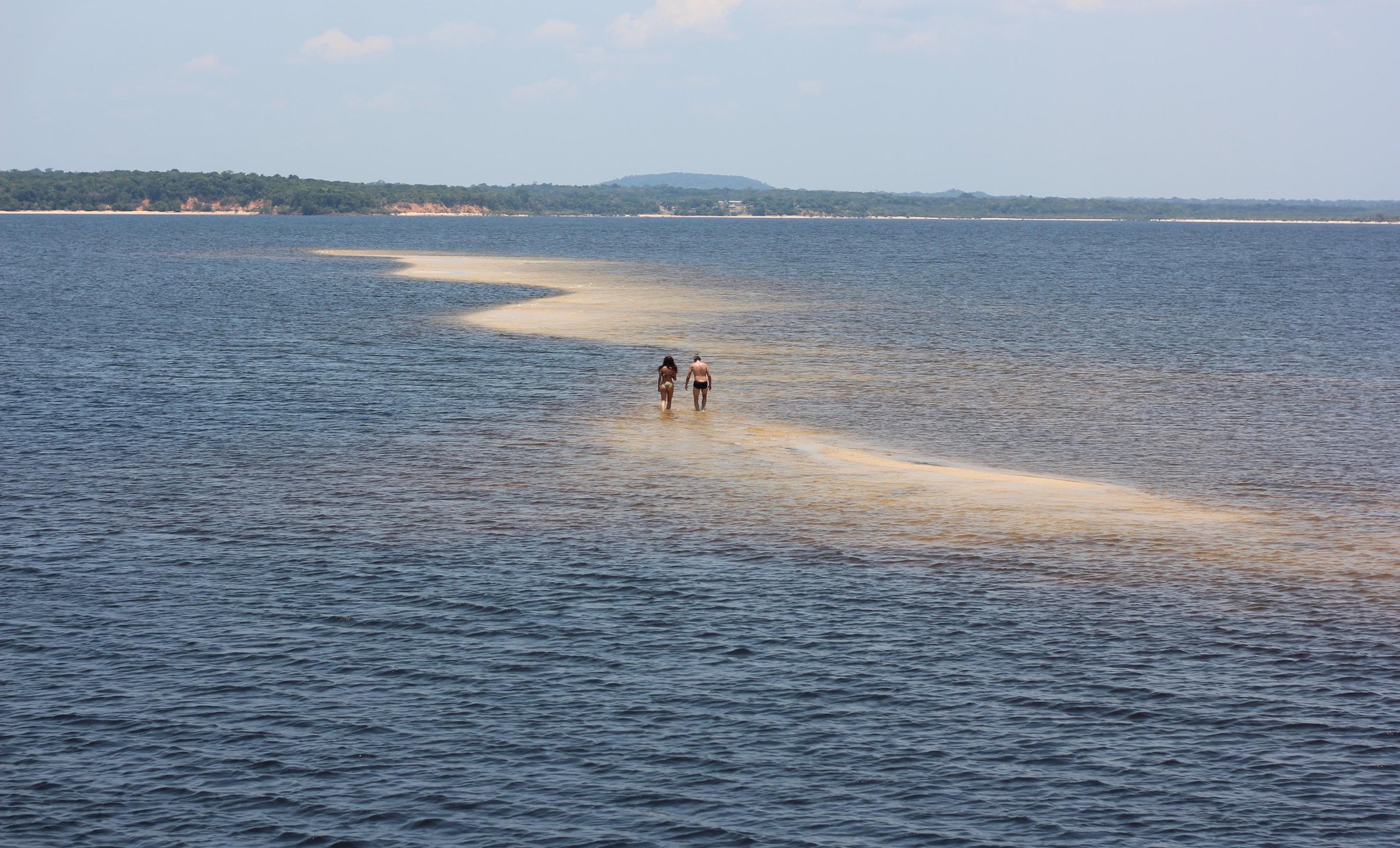 Eine Sandbank, die der Fluss nur gelegentlich freigibt. Zwei Reisende waten entlang einer schmalen Sandbank, die sich über breite blaue Flusswasser erstreckt, auf einer Amazonas-Kreuzfahrt ab Alter do Chão, mit bewaldetem Ufer und sanften Hügeln im Hintergrund