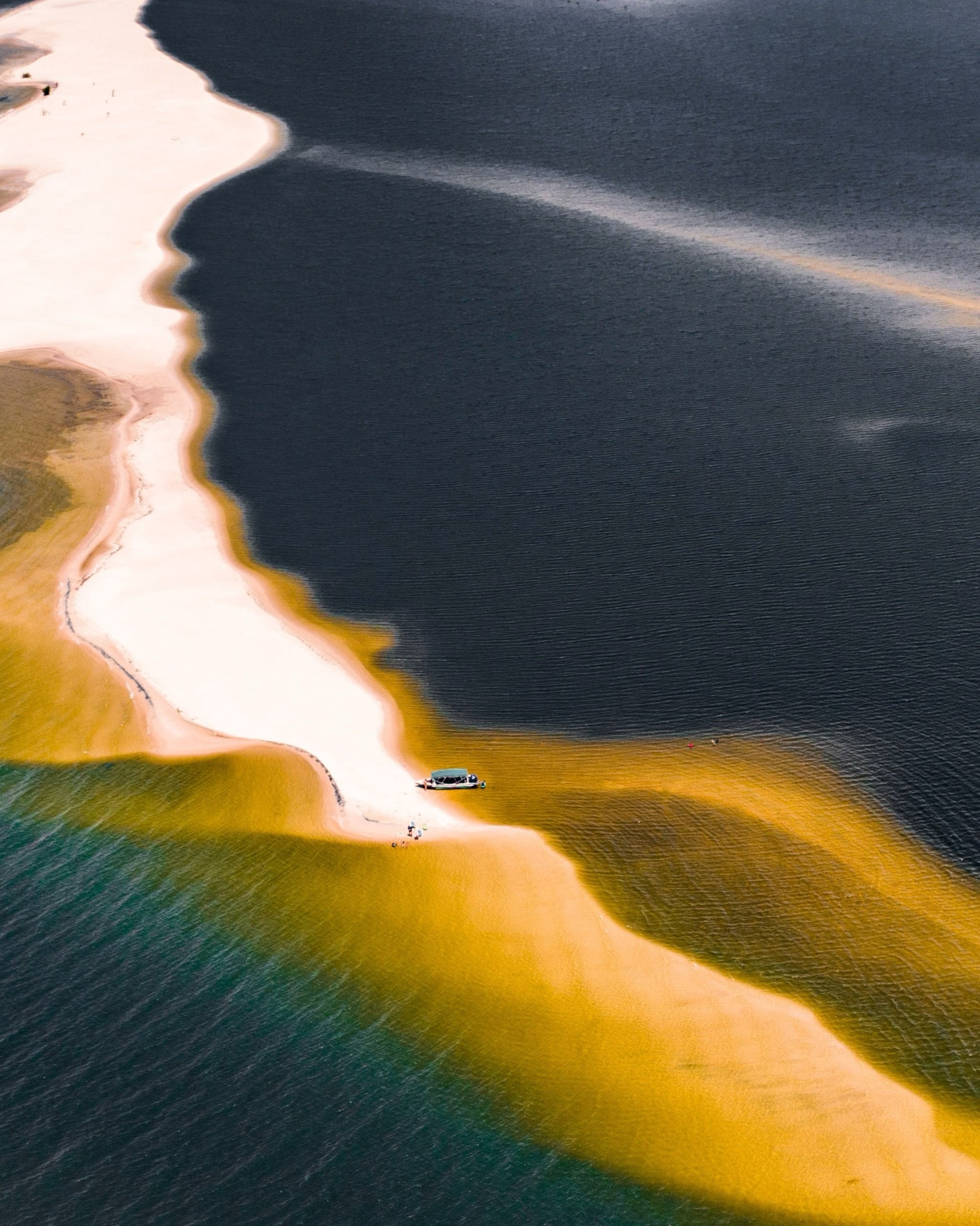 Sand, water, and three shades of river — a boat barely visible from above. Aerial view of a small boat moored on a winding white sandbar where teal, gold and dark river waters meet during an Amazon cruise from Alter do Chão