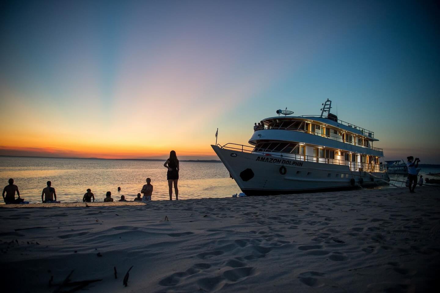 Guests relaxing on a white sand river beach at sunset beside the Amazon Dolphin vessel, part of an Amazon cruise from Alter do Chão, with a vivid orange and blue sky reflected on calm water