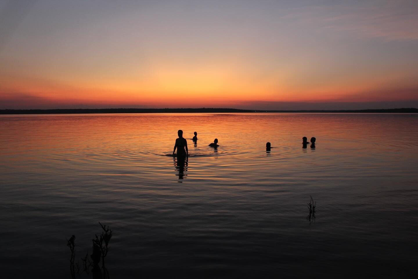 Warm river, burning sky — the perfect end to a day on the water. Silhouettes of travellers swimming in a vast, glassy river at dusk on an Amazon cruise from Alter do Chão, with a fiery orange and red sunset reflected across the water