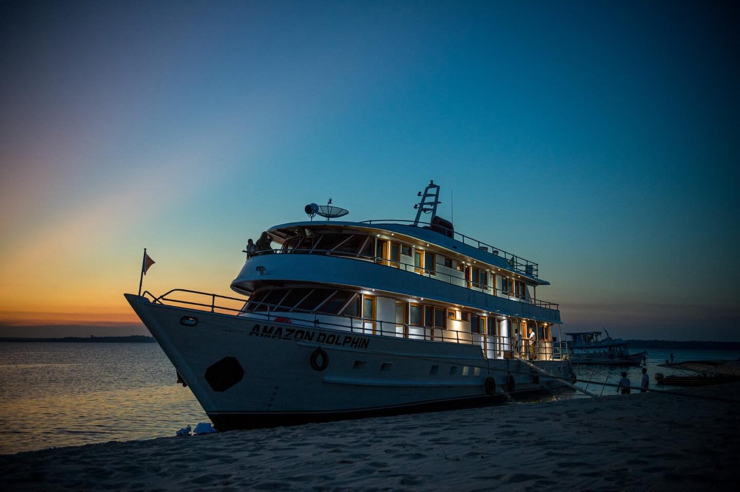 Das schwimmende Lager wartet, bevor der Fluss in die Nacht taucht. Das Kreuzfahrtschiff Amazon Dolphin liegt bei Einbruch der Dämmerung an einer Sandbank, die warmen Kabinenlichter leuchten vor einem tiefblauen und orangefarbenen Abendhimmel
