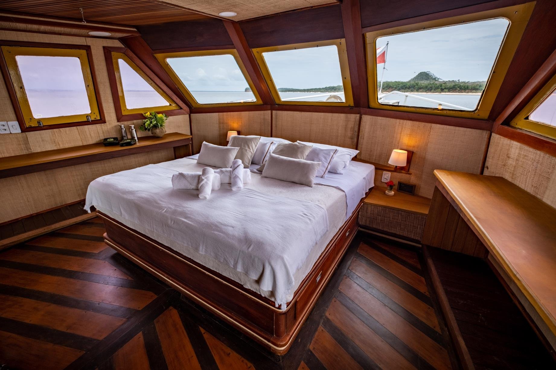 Jeden Morgen ein neues Panorama, direkt vom Bett aus. Geräumige Hauptkabine auf einer Amazonas-Kreuzfahrt ab Alter do Chão mit Kingsize-Bett in weißer Bettwäsche, gefalteten Handtuchtieren, warmem Holzdekor und Panoramafenstern mit Blick auf ein bewaldetes Flussufer