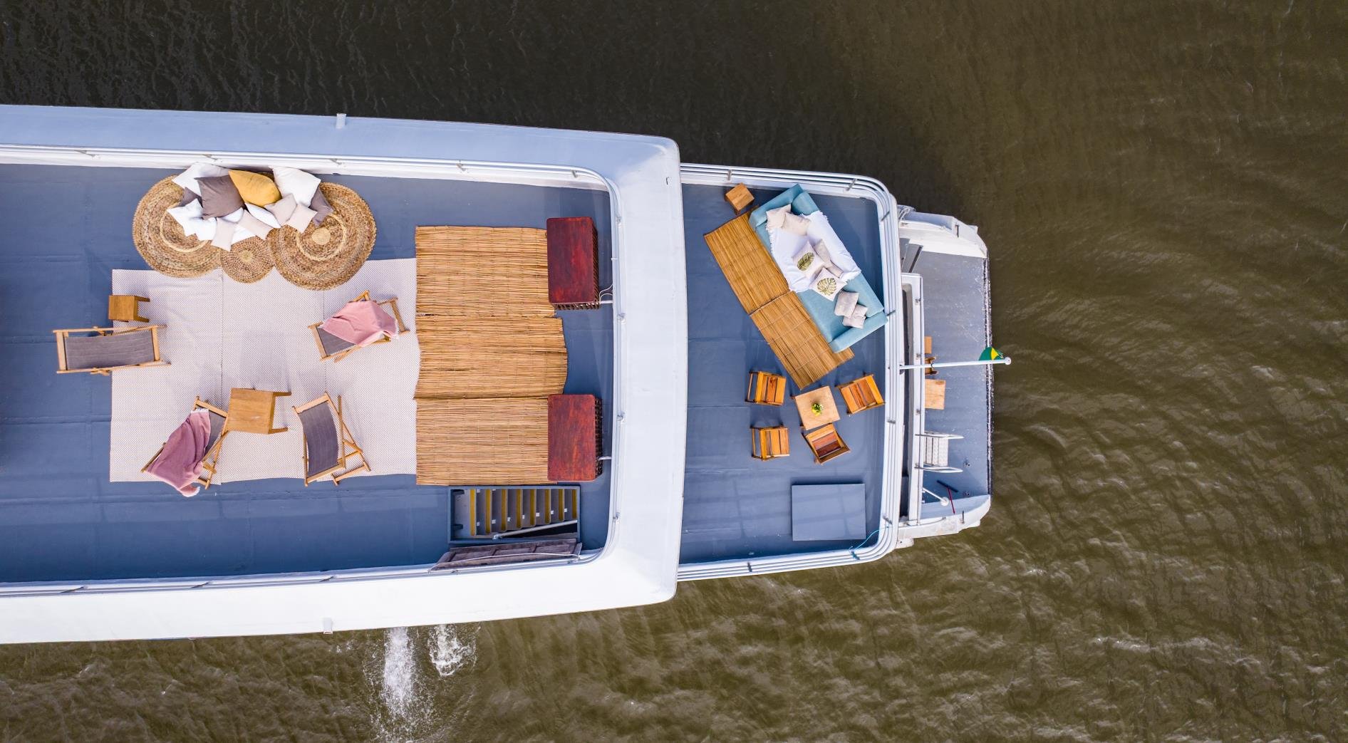Every centimetre of open deck put to work — wooden loungers, rattan accents, thatched shade and cushioned sofas, all moving slowly upriver. Aerial view of a luxury Amazon cruise from Alter do Chão showing two open sundecks furnished with wooden loungers, rattan rugs, thatched shade structures and cushioned seating, cruising across a wide brown river