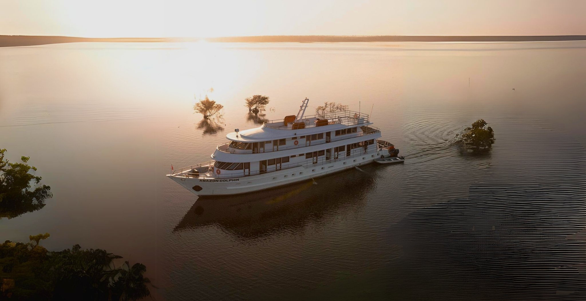 The Amazon Dolphin vessel gliding across a vast flooded river at golden sunset on an Amazon cruise from Alter do Chão, surrounded by partially submerged trees and mirror-calm water