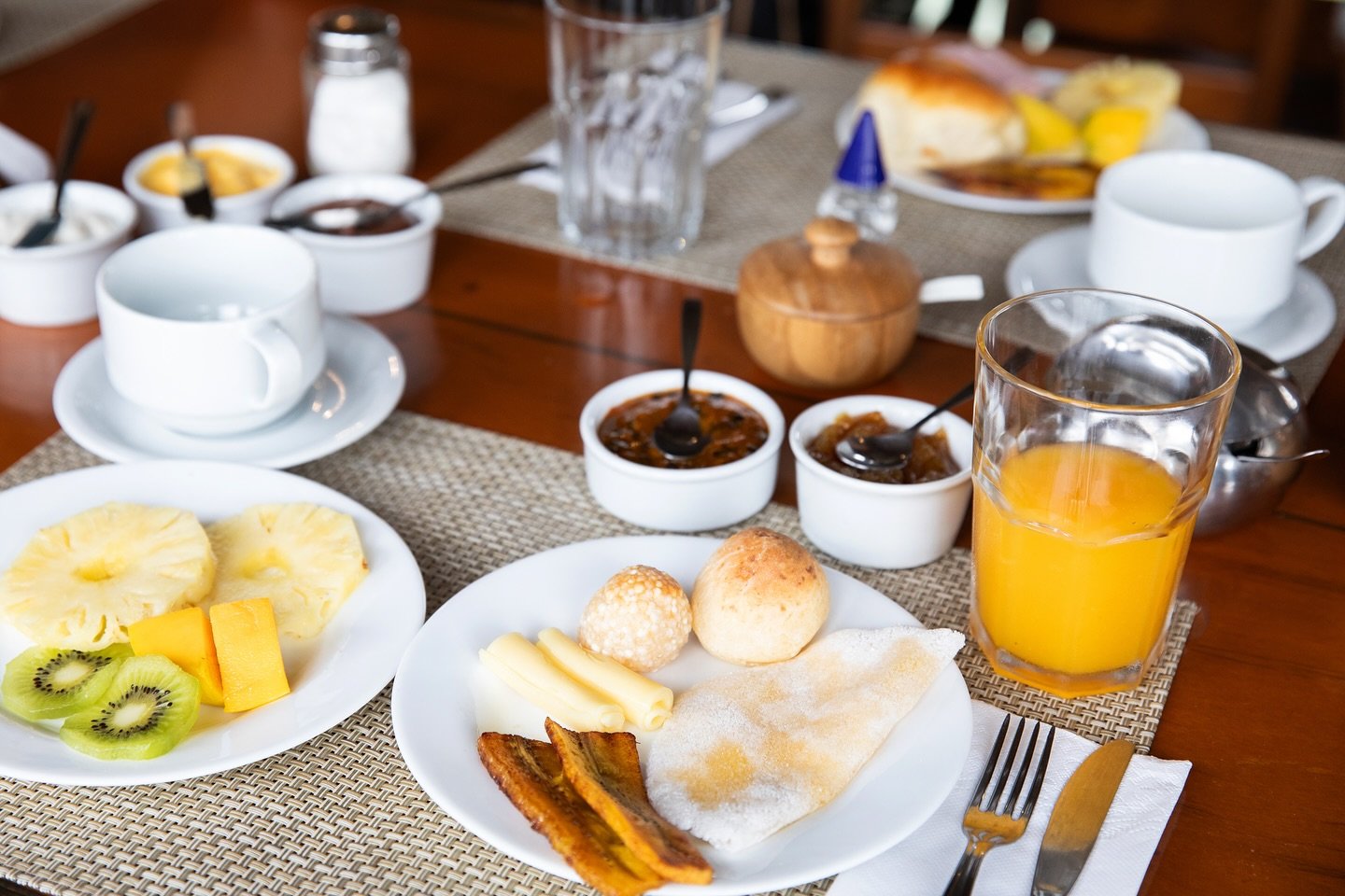 Tropical fruits, tapioca, and cheese rolls — fuel for a full day on the river. A generous Amazon Cruise Trip in Brazil breakfast laid out on a wooden table, featuring pão de queijo, rolled cheese slices, fried plantains, a tapioca crepe, fresh pineapple and kiwi, orange juice, and small bowls of jam alongside coffee cups