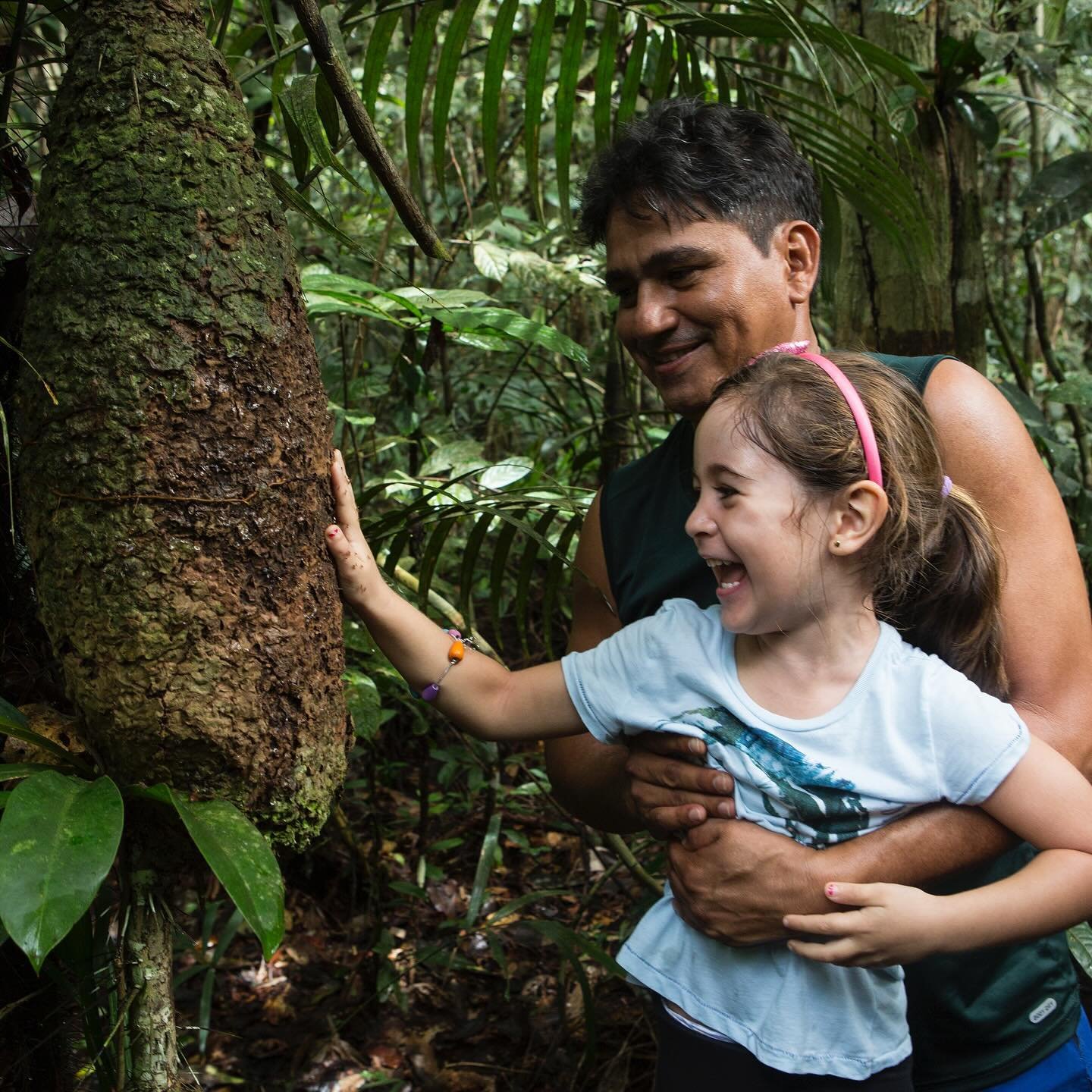 Pure delight — fingertips on ancient bark, forest all around. A laughing girl touches the textured bark of a large rainforest tree during an Amazon Cruise Trip in Brazil, held gently by a smiling adult guide behind her