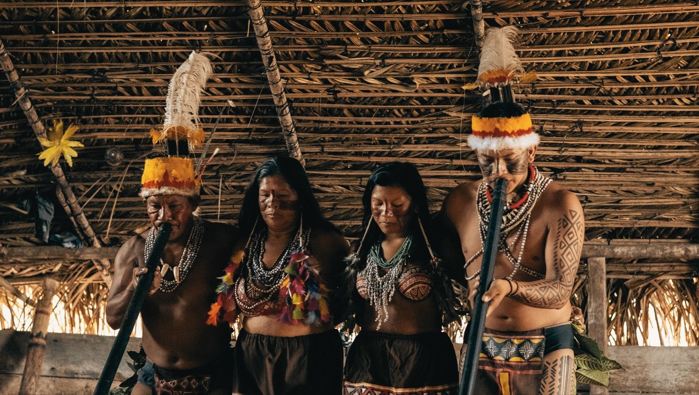 Feathers, geometric tattoos, and centuries of living tradition — all under one thatched roof. Four Indigenous people in traditional feathered headdresses, body paint, and layered bead necklaces performing a ritual inside a thatched-roof longhouse during an Amazon Cruise Trip in Brazil
