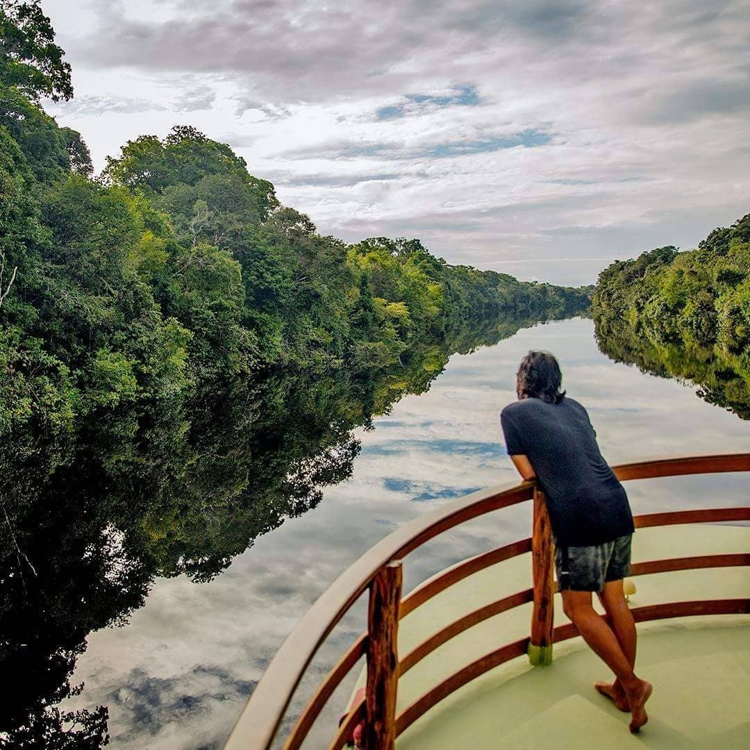 Água de espelho, selva sem fim — o canal se abre à frente. Um viajante apoia-se no corrimão de madeira da proa de um barco fluvial e contempla um canal de floresta com águas perfeitamente espelhadas, ladeado por uma densa copa de floresta tropical, durante um cruzeiro pela selva amazônica no Brasil