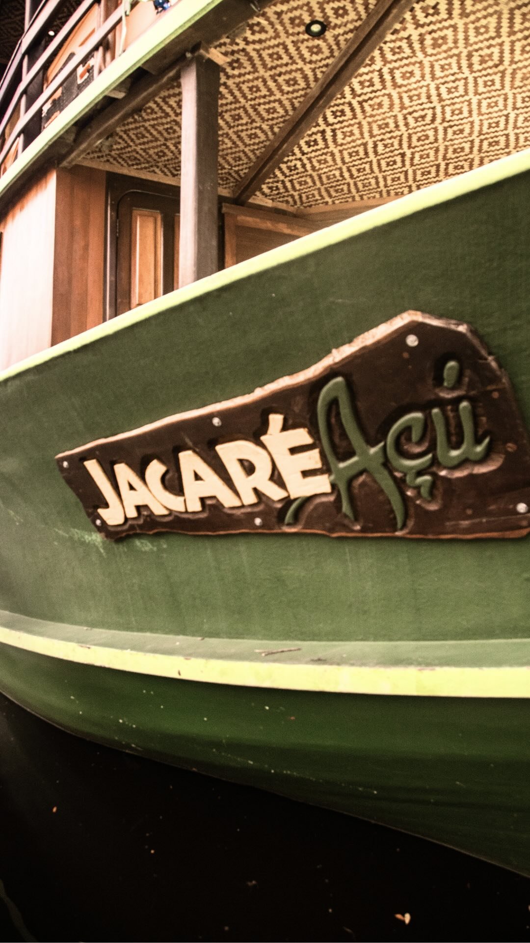Detalhe da placa de madeira esculpida com o nome 'Jacaré Açu' fixada no casco verde do barco de cruzeiro pela Amazônia brasileira, com teto estampado e estrutura de madeira ao fundo