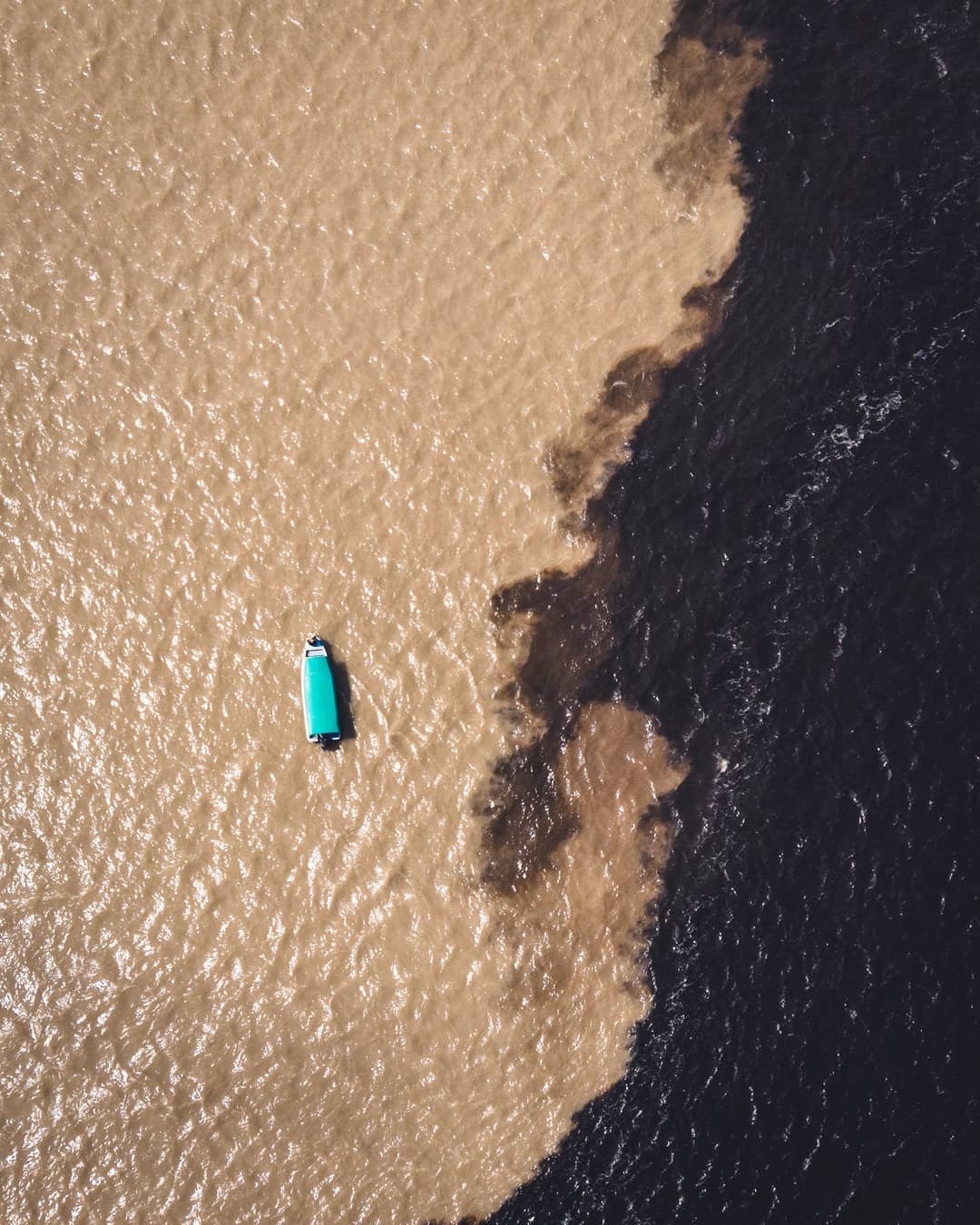 Dois rios lado a lado, sem se misturar. Vista aérea de um pequeno barco com teto turquesa em um cruzeiro pela Amazônia no Brasil, cruzando a fronteira entre a água cor de areia e a água preta no Encontro das Águas