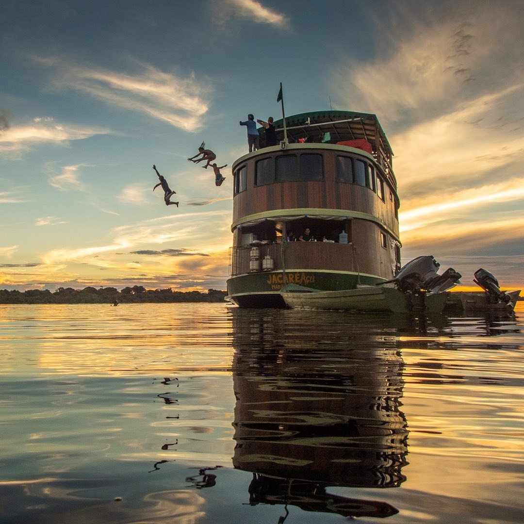 Pulo no espelho d'água — o fim de tarde que não se esquece. Passageiros pulando do convés superior do barco Jacaré Açu em um rio espelhado ao pôr do sol dourado durante um cruzeiro pela selva amazônica no Brasil