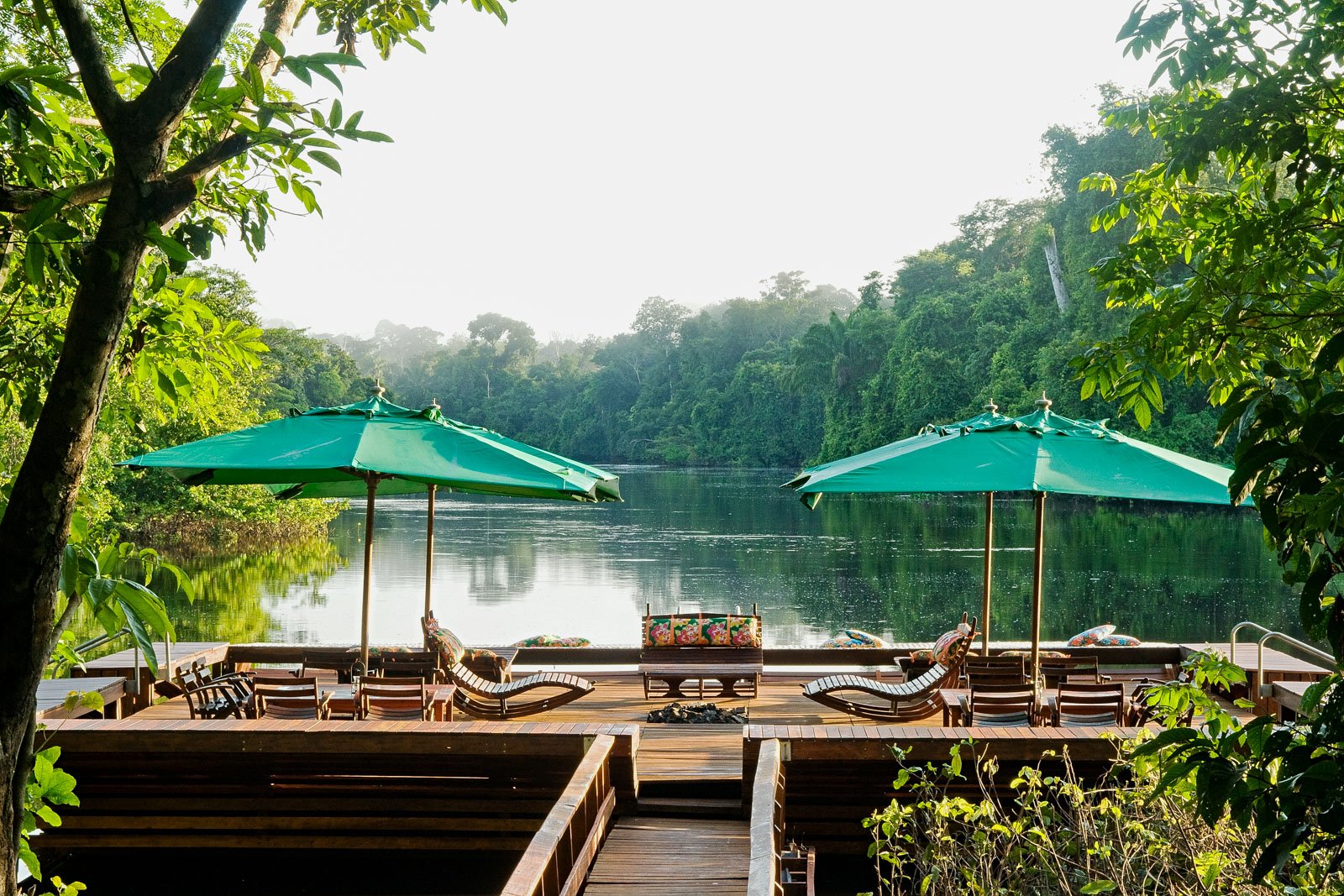 Wooden deck at an amazon jungle lodge in Brazil with teal parasols and rocking chairs overlooking a calm jungle river flanked by dense rainforest canopy
