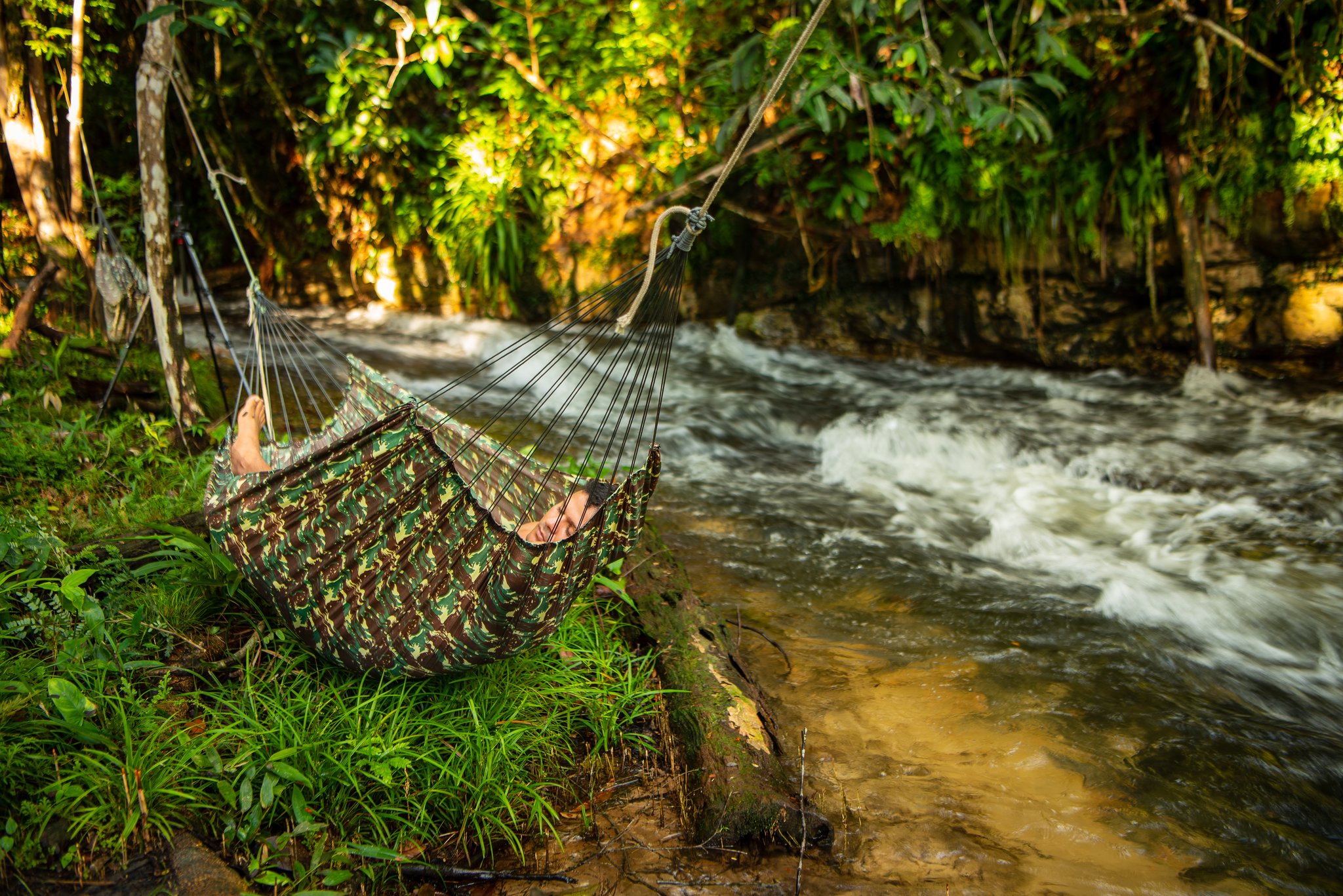 Stroom, boomschaduw en een hangmat: het enige antwoord op een lange peddeltocht. Een reiziger rust in een camouflage hangmat die naast een kolkende junglestroom hangt, omgeven door weelderig tropisch groen tijdens een Amazon kajakrondvaart in Brazilië
