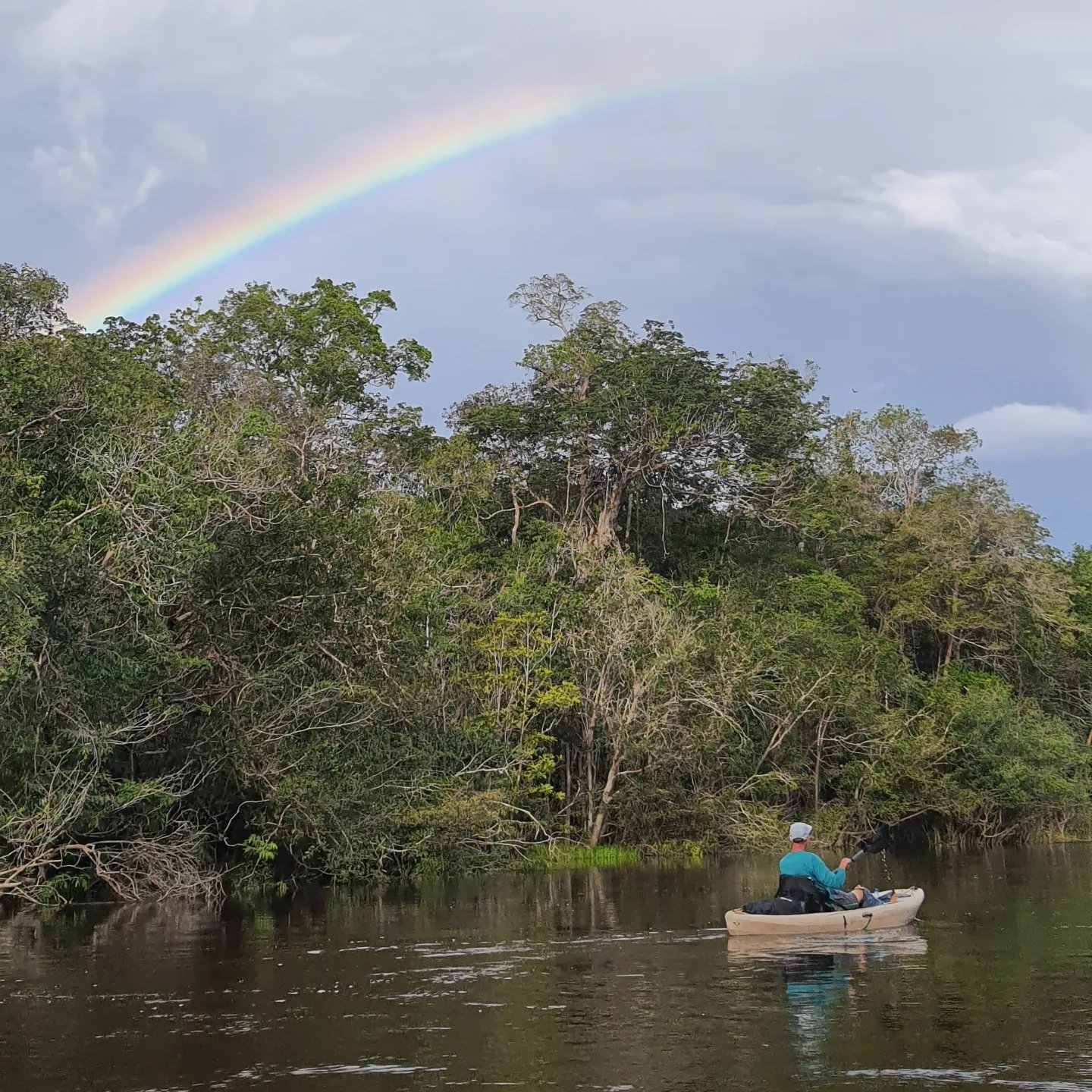 Soms peddel je gewoon de Amazone in en verschijnt er een regenboog. Een solo-kajakker peddelt over een donkerwater rivier langs dicht regenwoud terwijl een heldere regenboog zich uitstrekt over de bewolkte lucht tijdens een Amazon-kajaktrip in Brazilië
