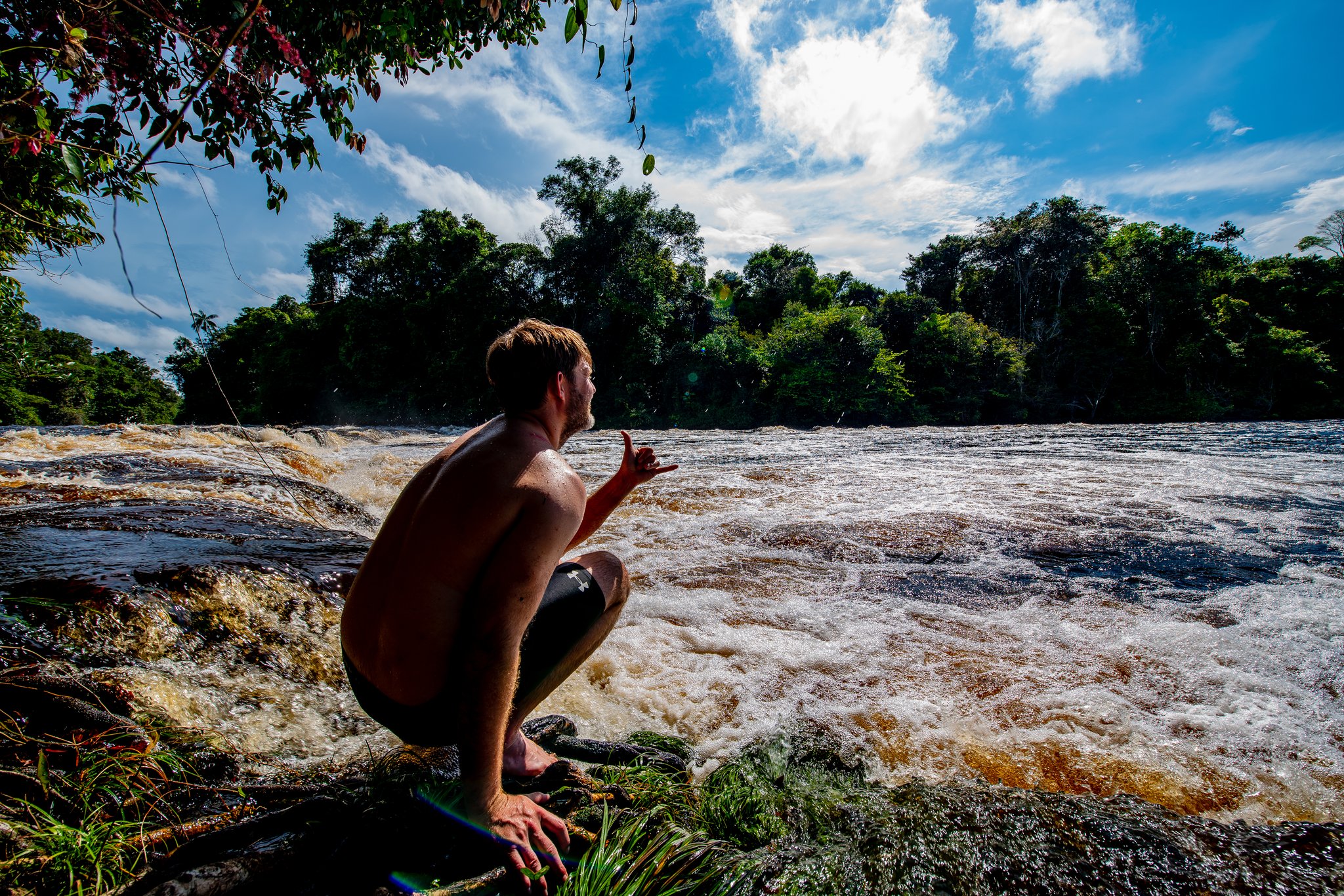 Even de stroming inschatten, voor die dat zelf doet. Een reiziger gehurkt op de rotsachtige rand van bruisende stroomversnellingen tijdens een Amazon-kajaktour in Brazilië, omgeven door dicht regenwoud onder een stralend blauwe lucht