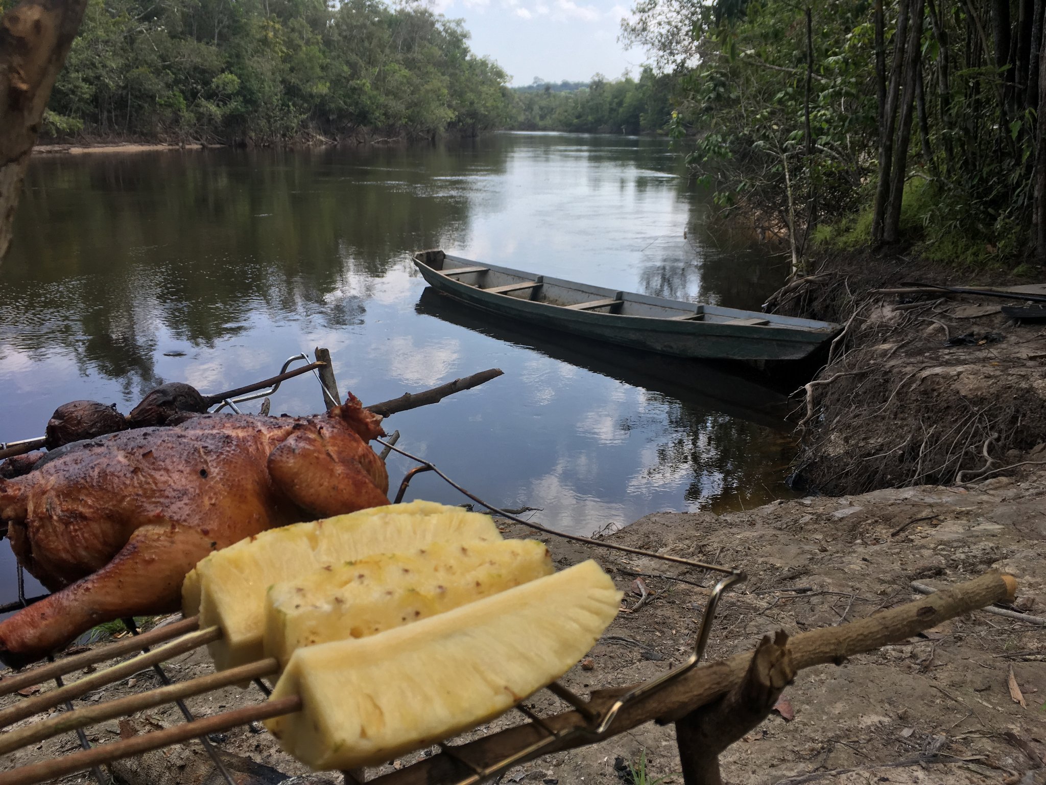 Barbecue aan het water, kano klaar voor vertrek, het oerwoud dicteert het tempo. Gegrild vlees en verse ananasschijven op bamboespiesen aan de rivieroever tijdens een Amazon-kayaktour in Brazilië, met een houten kano aangemeerd op een rustige junglerivier op de achtergrond