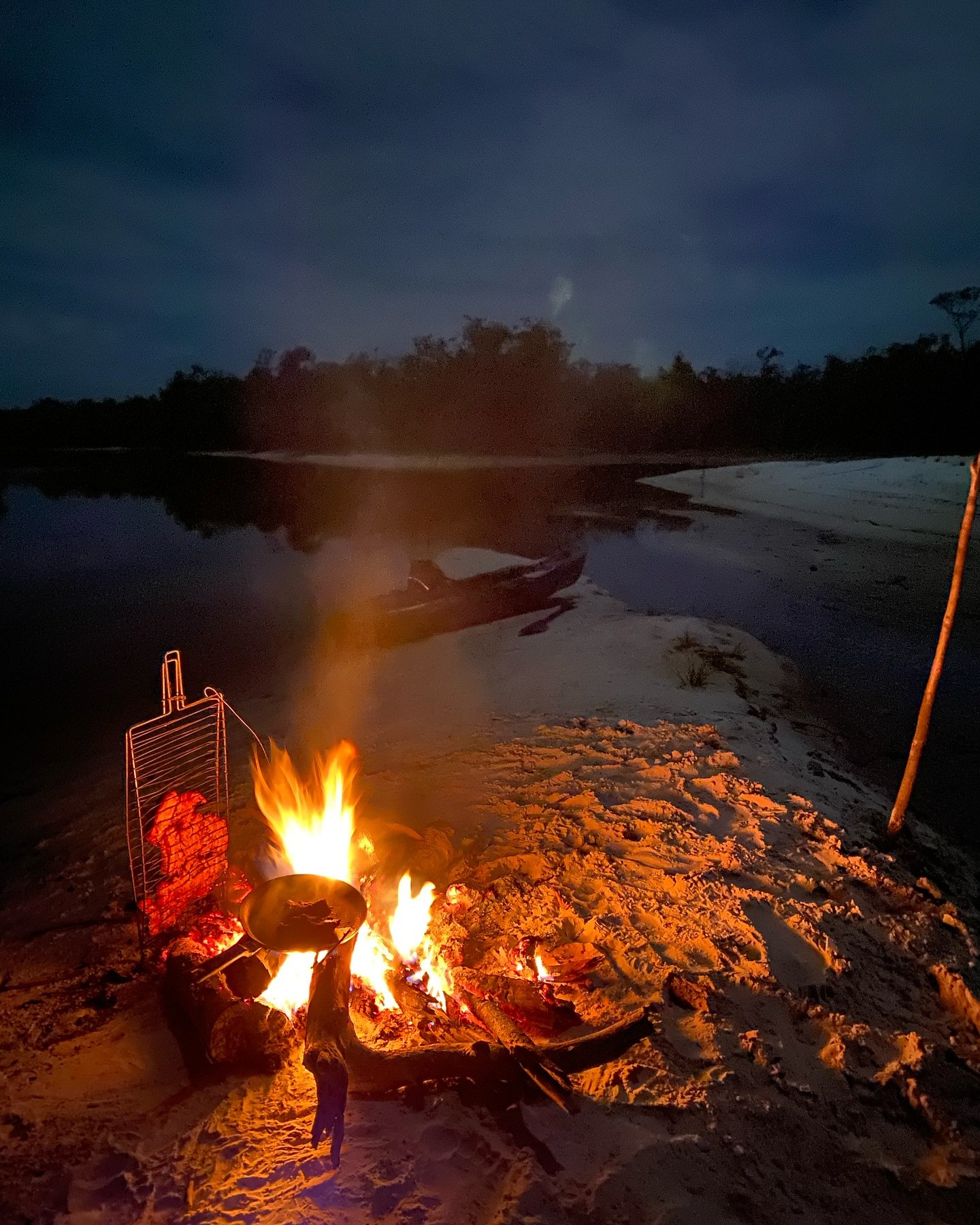 Boot vast, vuur aan, pan op. De Amazone kent geen menu's. Een knisperend kampvuur met een kookpan en grillrooster op een wit zandstrand aan de rivier 's nachts, met een houten kano aan de waterkant tijdens een Amazon-kajaktour in Brazilië
